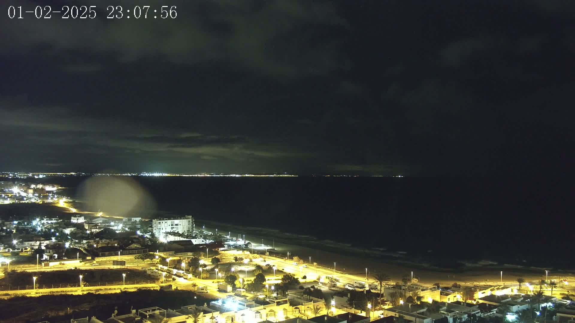 A nighttime coastal town with illuminated streets and buildings overlooks a dark ocean under a cloudy sky.