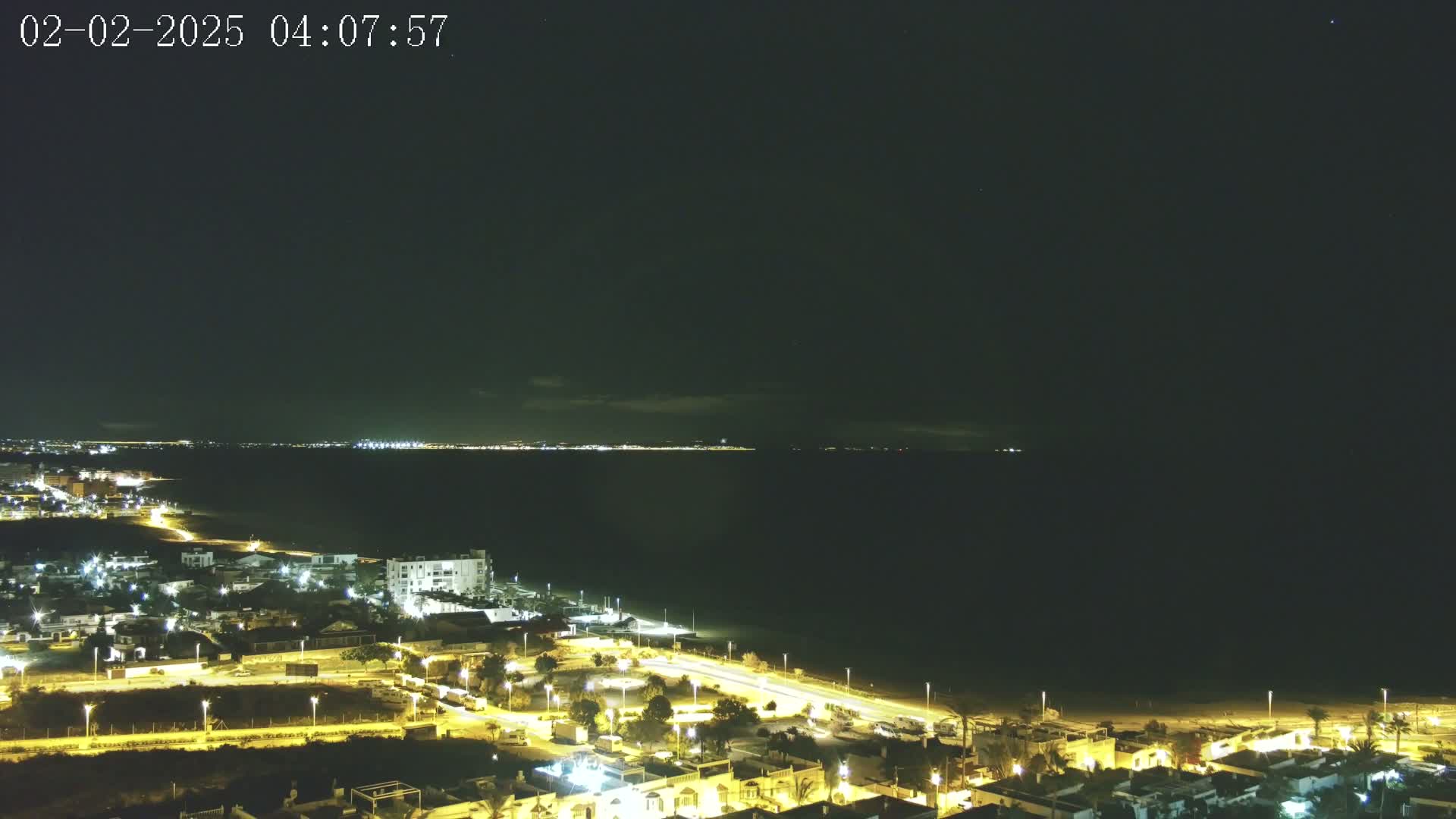 A nighttime coastal scene shows a dark ocean, a dimly lit beach, and a town with illuminated buildings and streets under a mostly clear, dark sky.