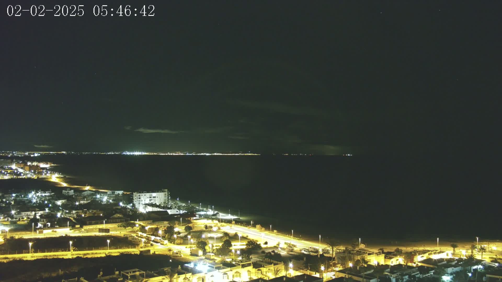 A nighttime coastal scene shows a dark ocean with a faintly lit cityscape on the horizon, and a brightly lit residential area next to a beach in the foreground under a mostly clear, dark sky.