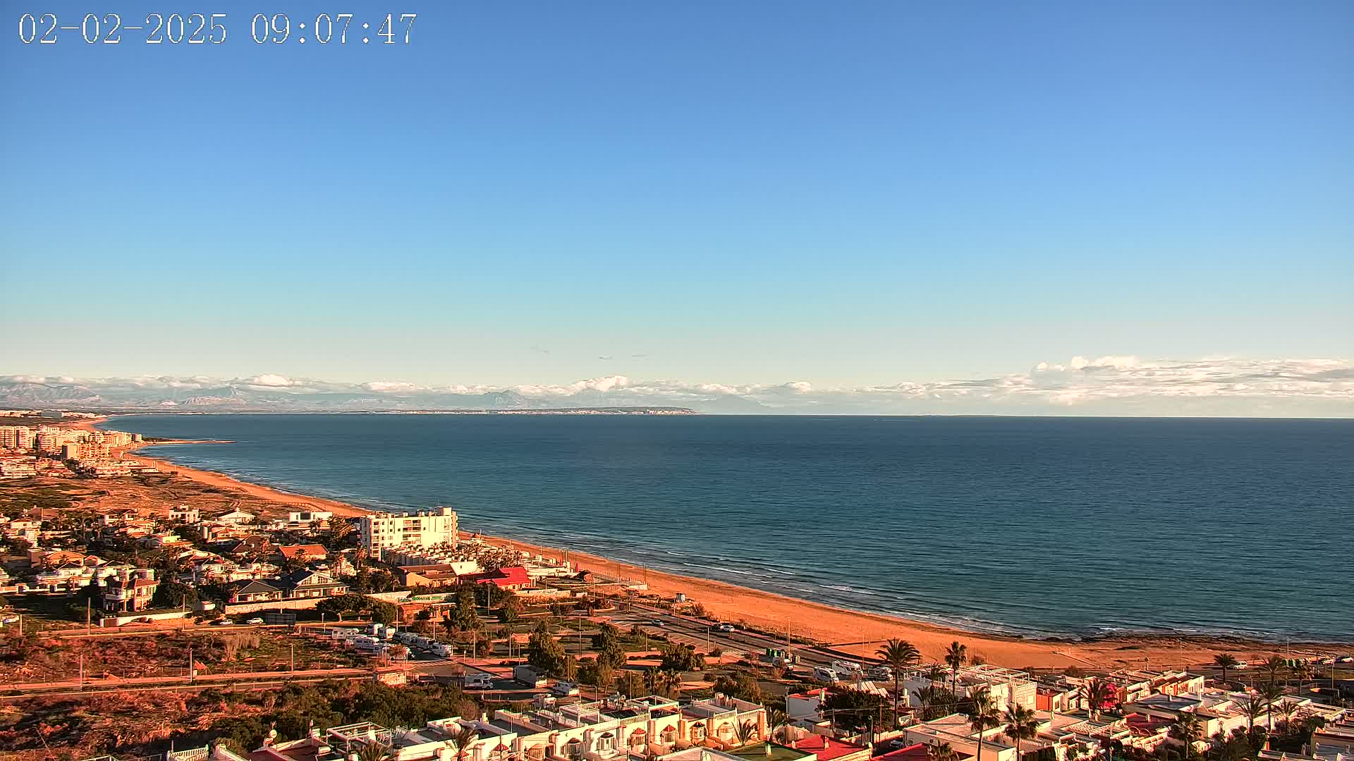 A coastal town with sandy beach and calm ocean under a mostly clear blue sky, mountains visible in the distance.
