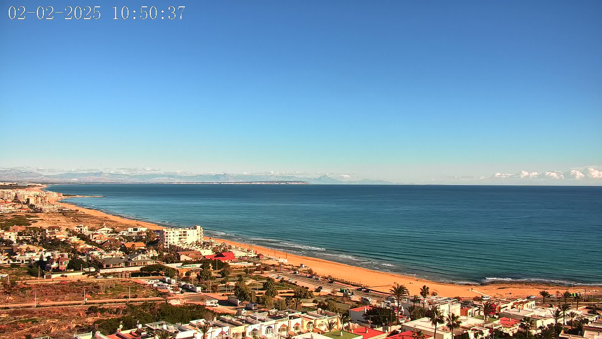 A coastal town overlooks a long, sandy beach and a calm ocean under a clear blue sky with a few distant clouds and mountains on the horizon.