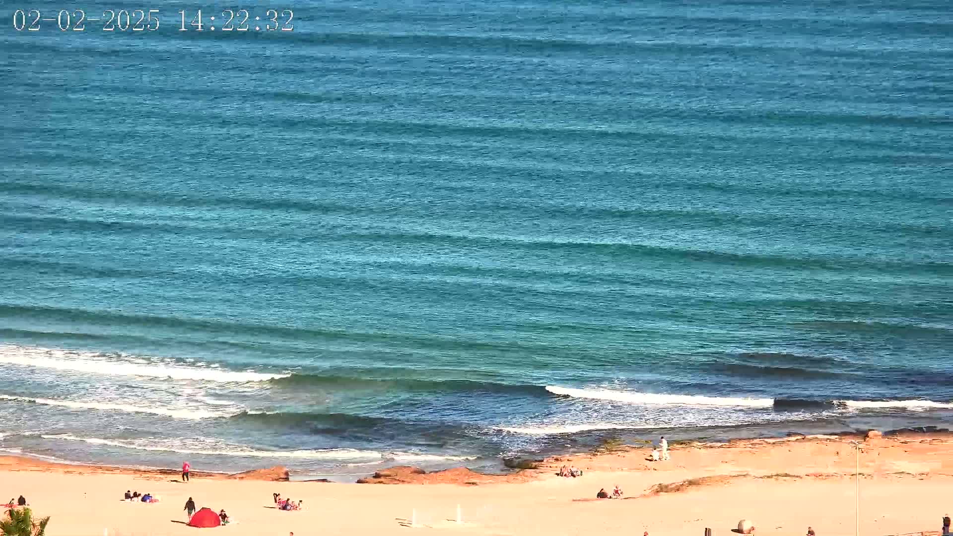 A sandy beach with a few people on it slopes down to a teal ocean with small waves under a sunny sky.