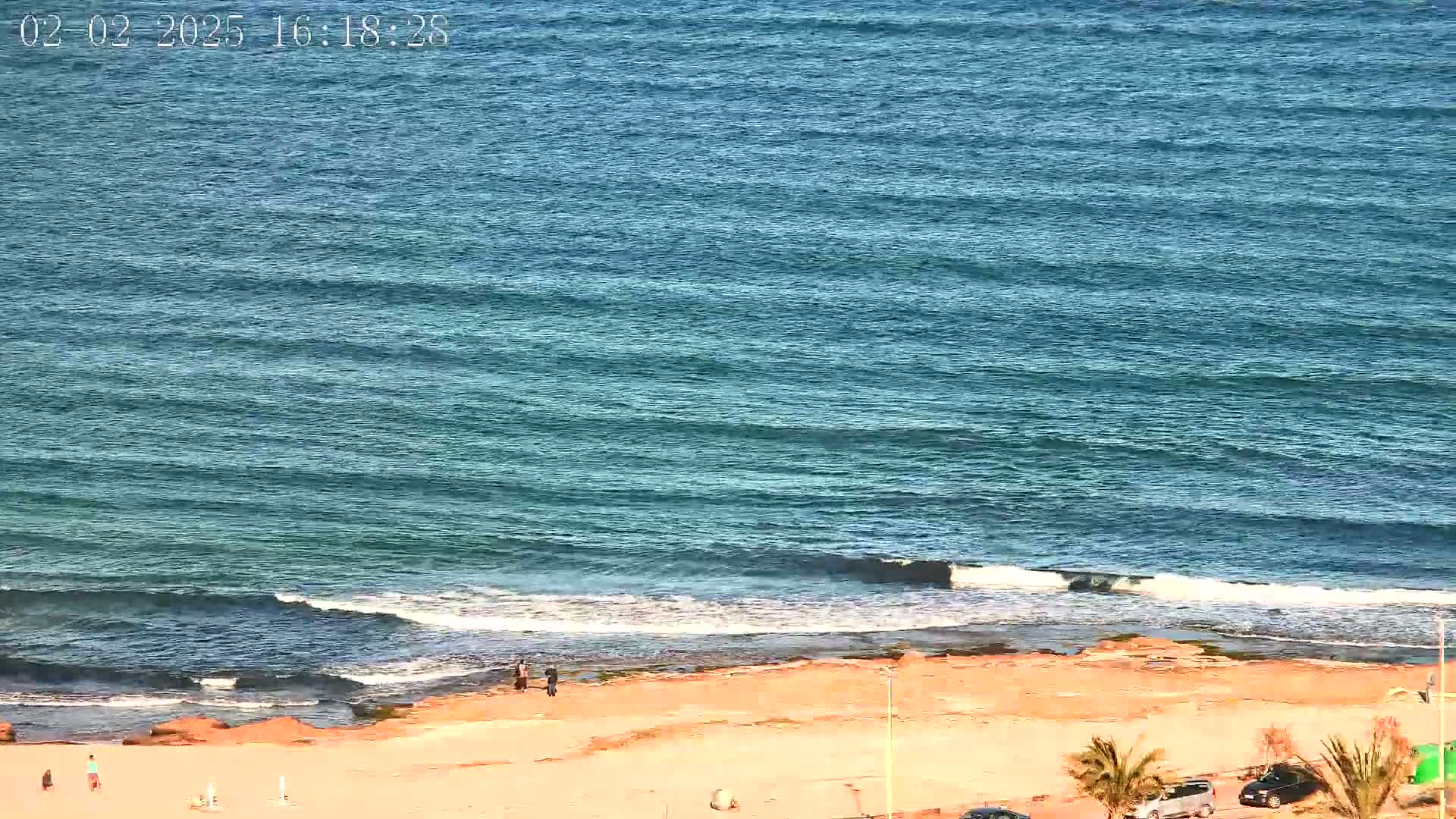 A beach with a few people on it is shown with gently rolling waves lapping at a sandy shore on a sunny day.
