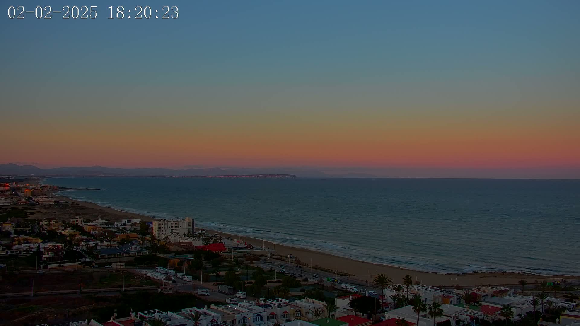 A coastal town at sunset overlooks a calm ocean with a pink and orange sky, and distant mountains are visible on the horizon.