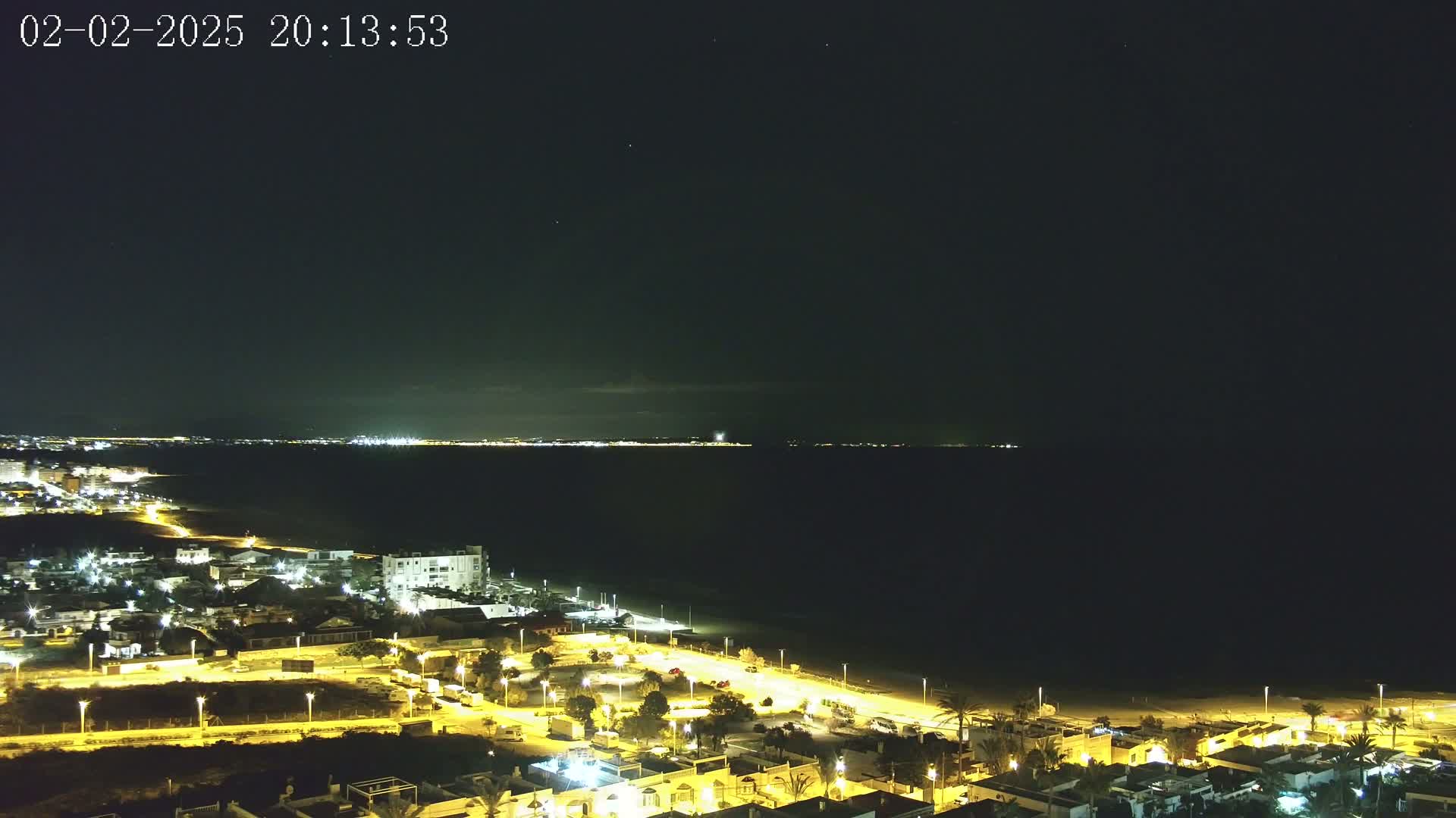 A night-time, aerial view of a coastal town illuminated by streetlights, overlooking a dark ocean under a clear, starry sky.