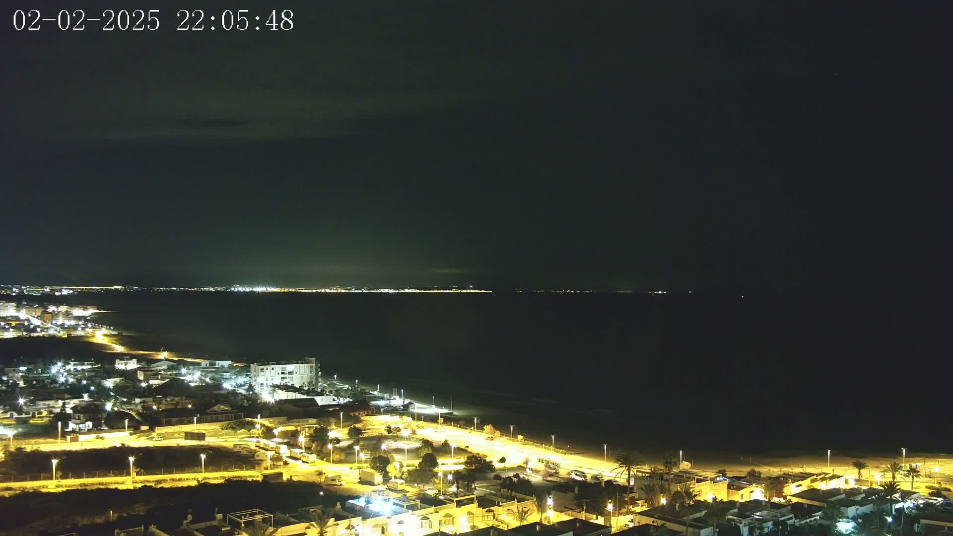 A nighttime coastal scene shows a dark ocean under a mostly clear sky, with a line of illuminated buildings along the shore.
