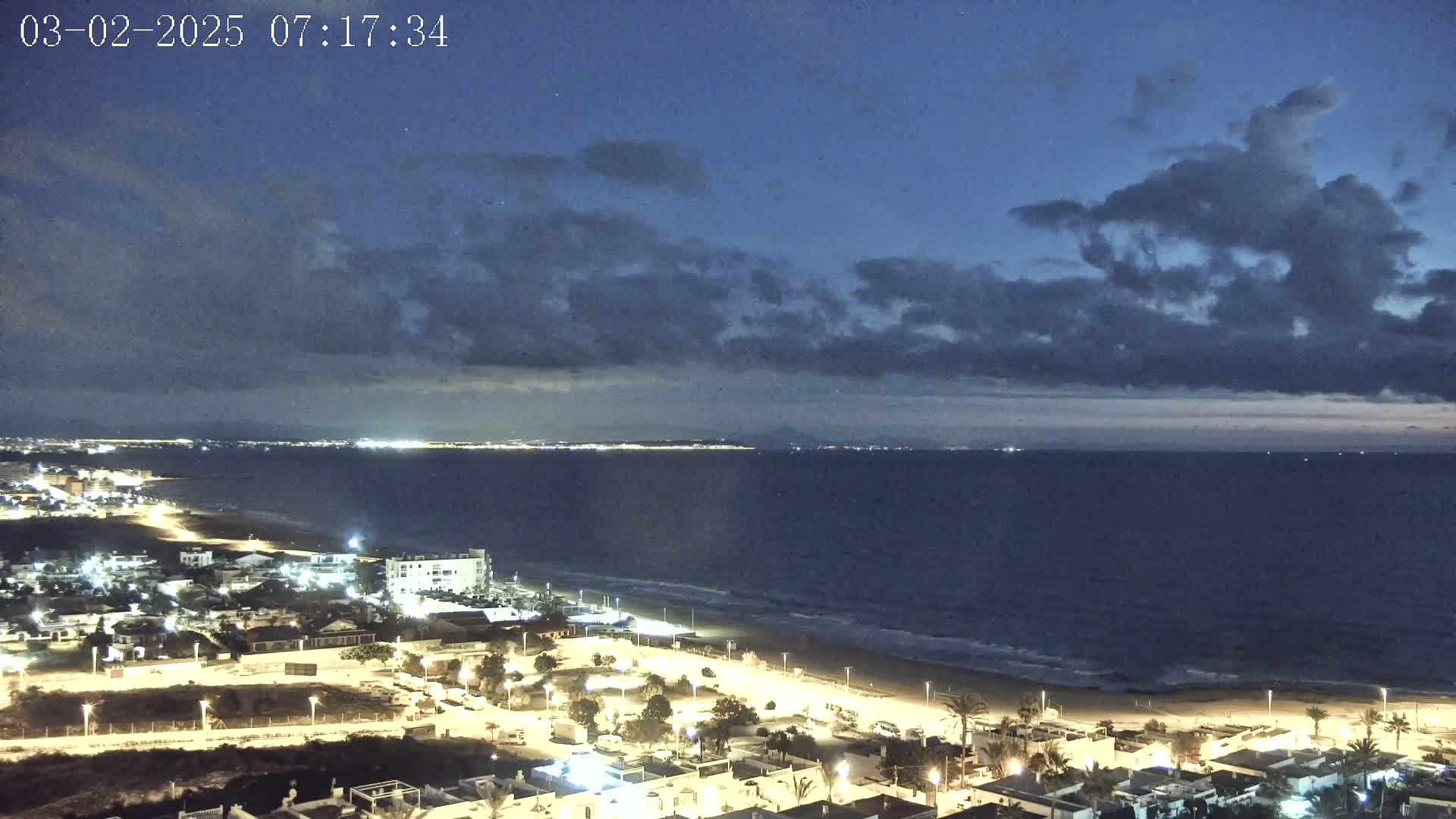 A nighttime coastal scene shows a dark, cloudy sky over a calm sea with a lit-up beach and town.