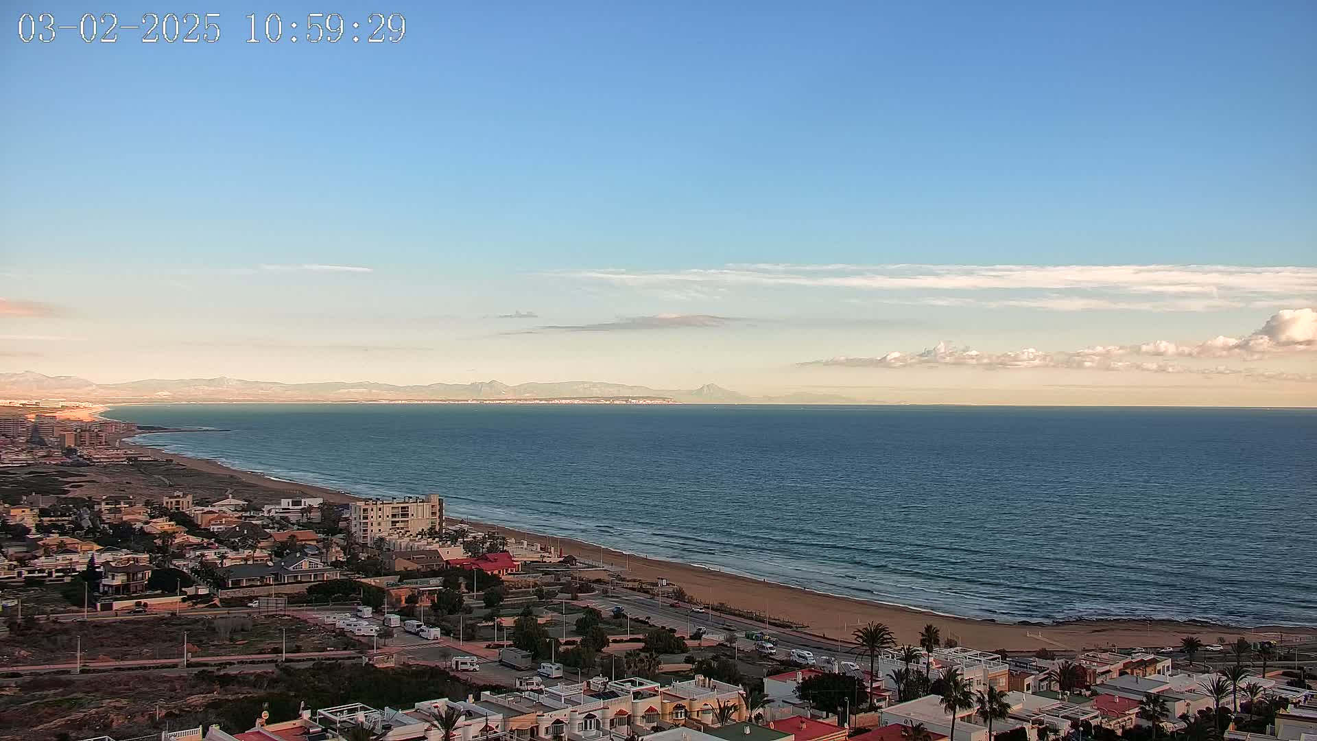 A coastal town overlooks a calm ocean under a mostly clear sky with a few scattered clouds, and distant mountains are visible on the horizon.