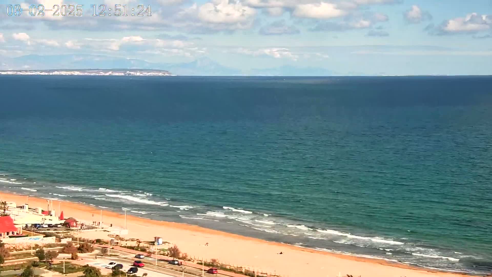 A sandy beach with gentle waves and a few people is visible, with a calm sea extending to a distant mountain range under a partly cloudy sky.