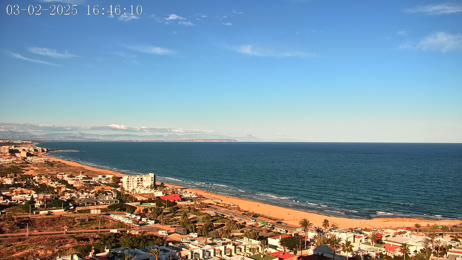 A coastal town overlooks a calm ocean under a mostly clear blue sky with distant mountains visible on the horizon.