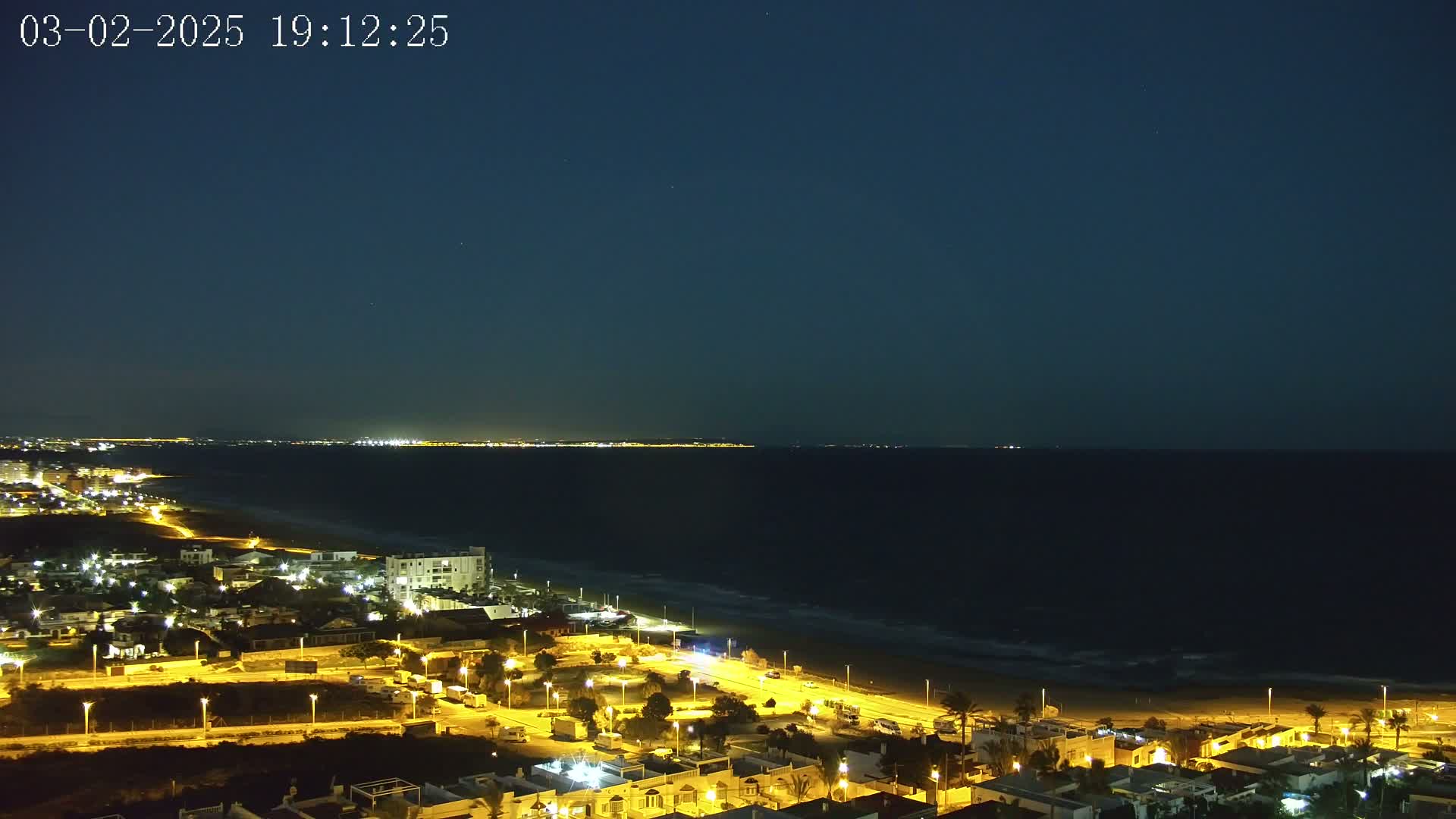 A nighttime coastal town is illuminated, with a dark ocean extending to a dimly lit distant shoreline under a clear, dark sky.
