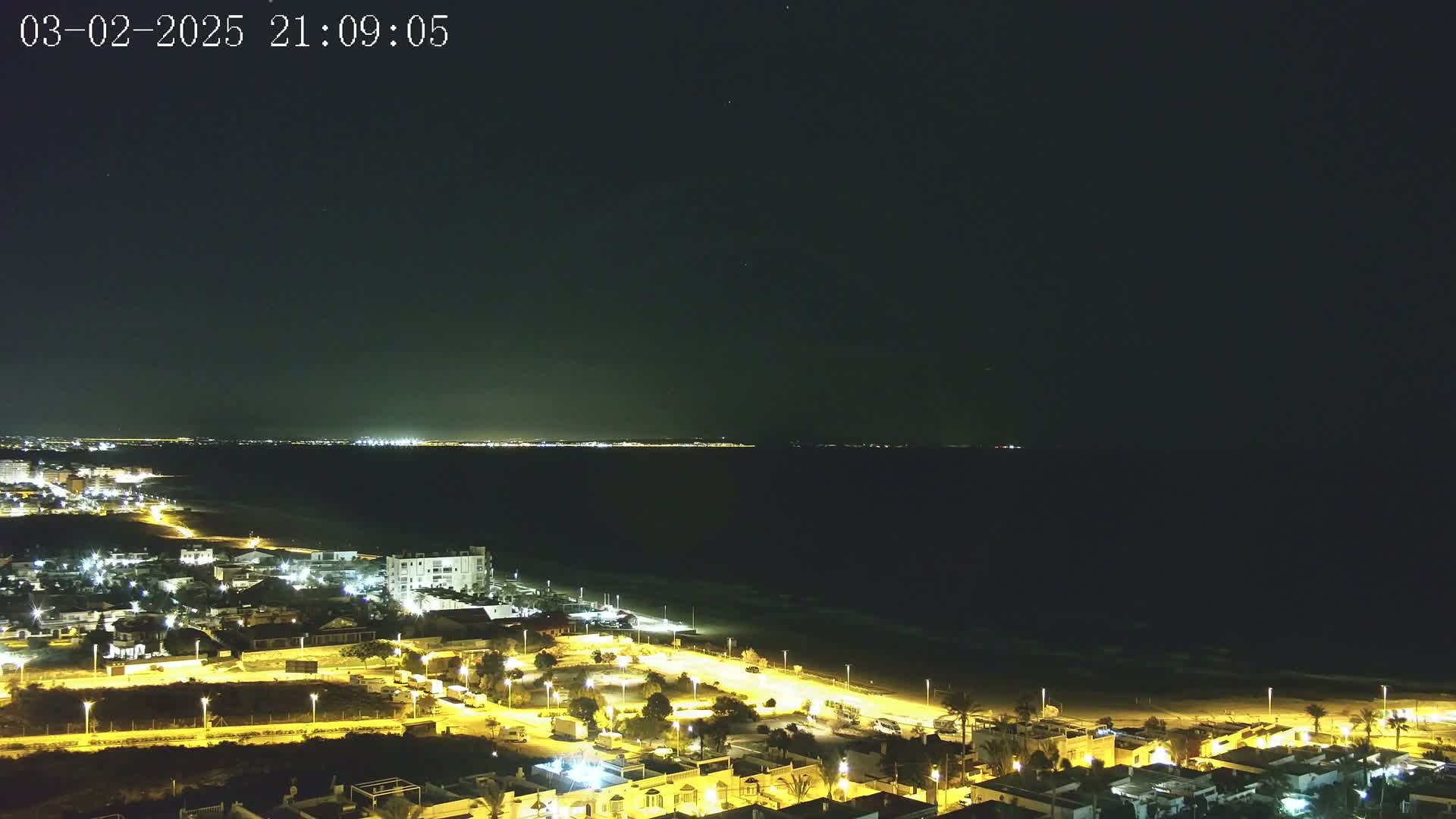 A nighttime aerial view shows a coastal town illuminated, with a dark ocean and a distant city skyline under a clear night sky.