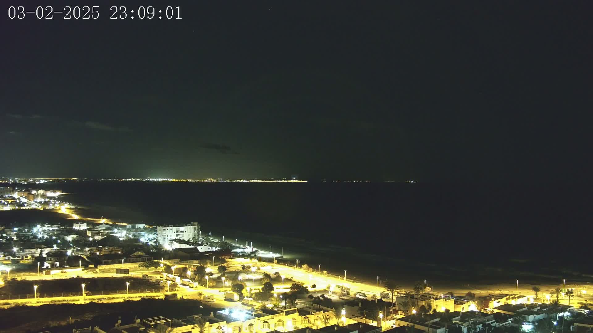 A nighttime aerial view shows a coastal town illuminated with streetlights, a dark beach, and a calm, dark sea extending to a distant, faintly lit shoreline under a mostly clear night sky.