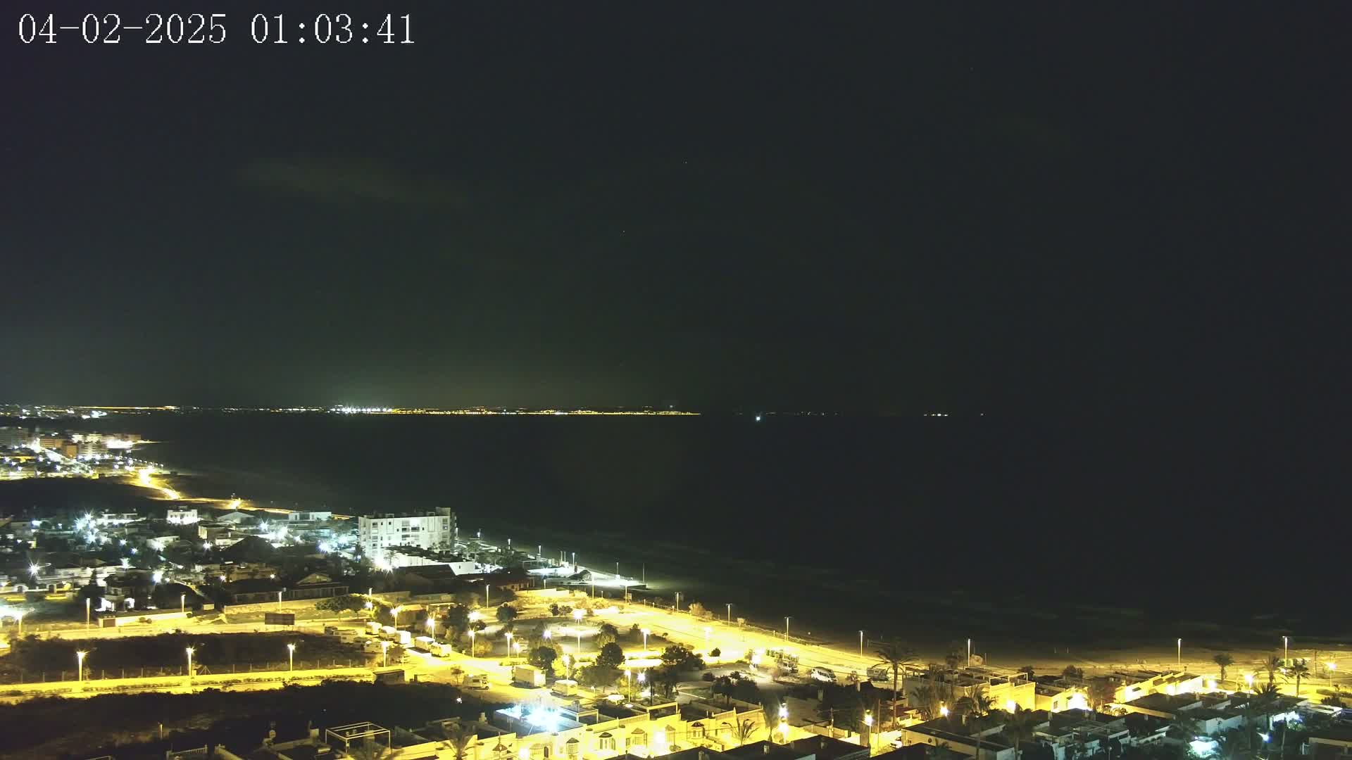 A nighttime coastal town with illuminated streets and buildings overlooks a dark ocean under a clear, starless sky.
