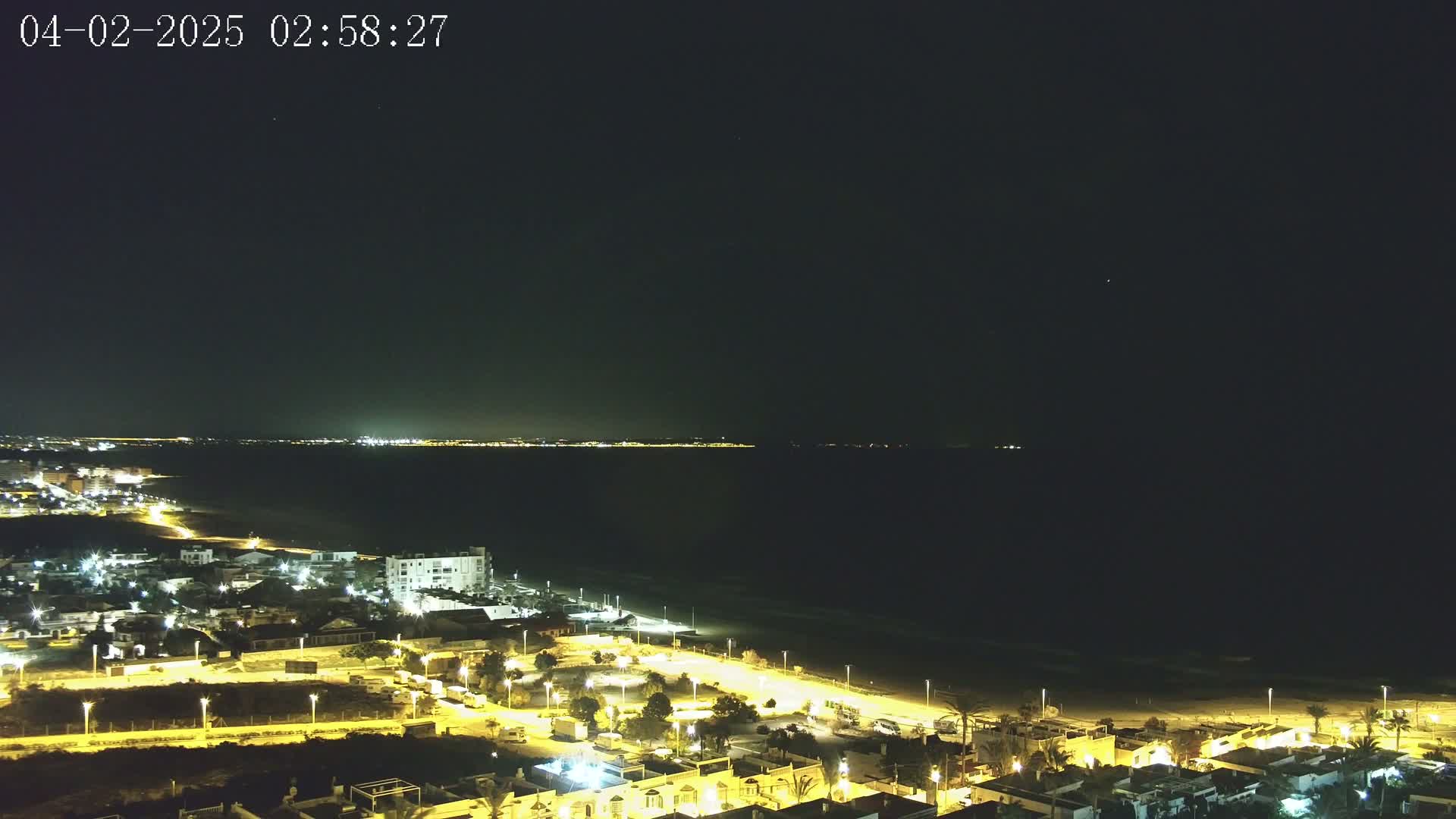 A night-time, aerial view shows a coastal town illuminated by streetlights, bordering a dark ocean with a faintly visible shoreline and distant city lights.