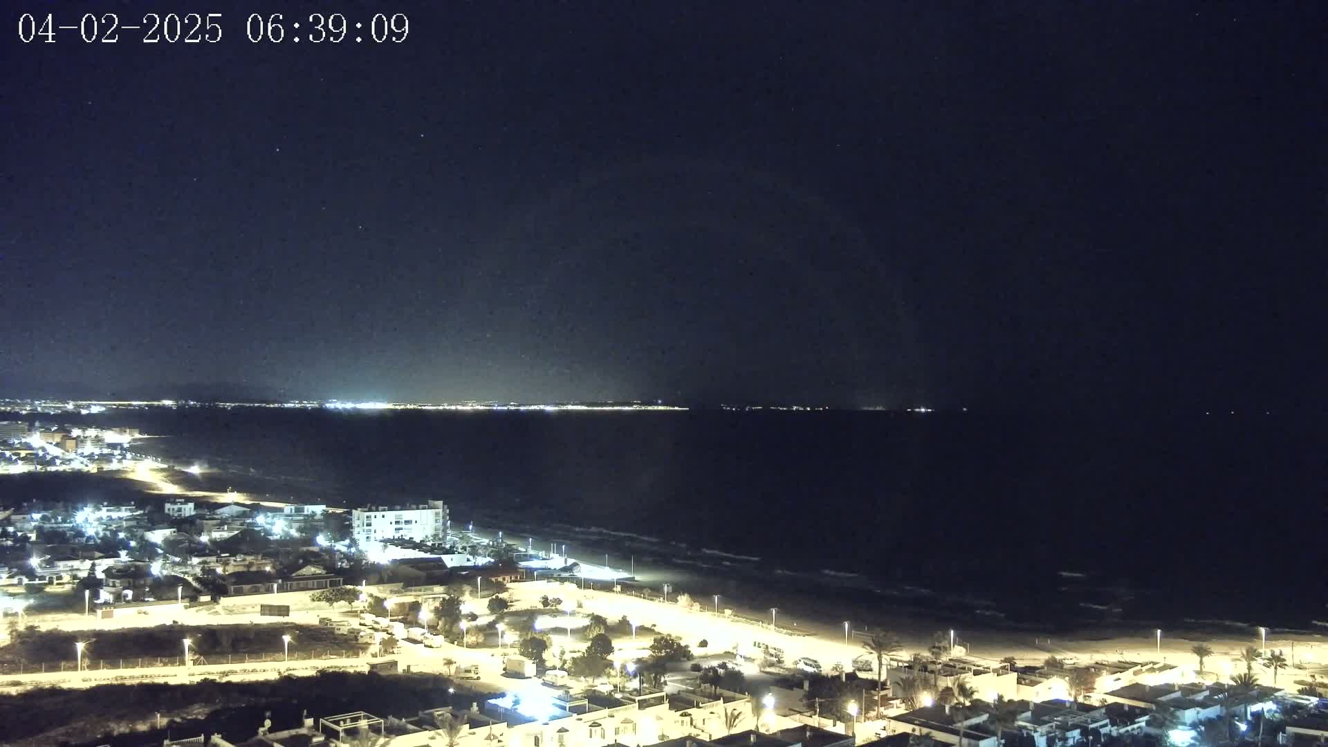 A nighttime coastal scene shows a dark ocean with small waves lapping a sandy beach, lined with illuminated buildings and streetlights, under a clear, starry sky.