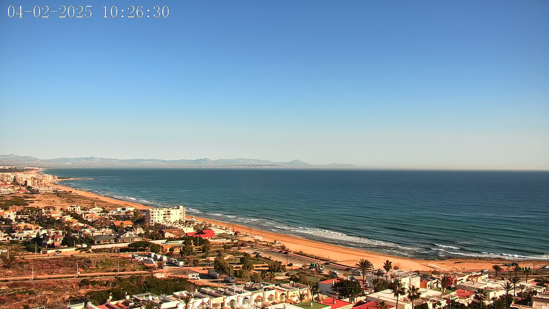 A coastal town overlooks a long sandy beach and a calm ocean under a clear blue sky, with mountains visible in the distance.