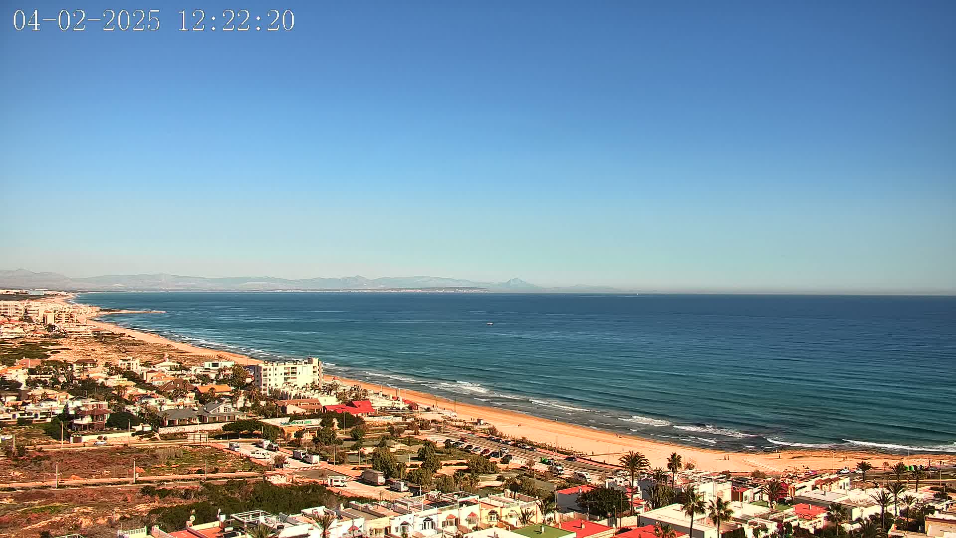 A coastal town overlooks a long, sandy beach and a calm ocean under a clear blue sky.