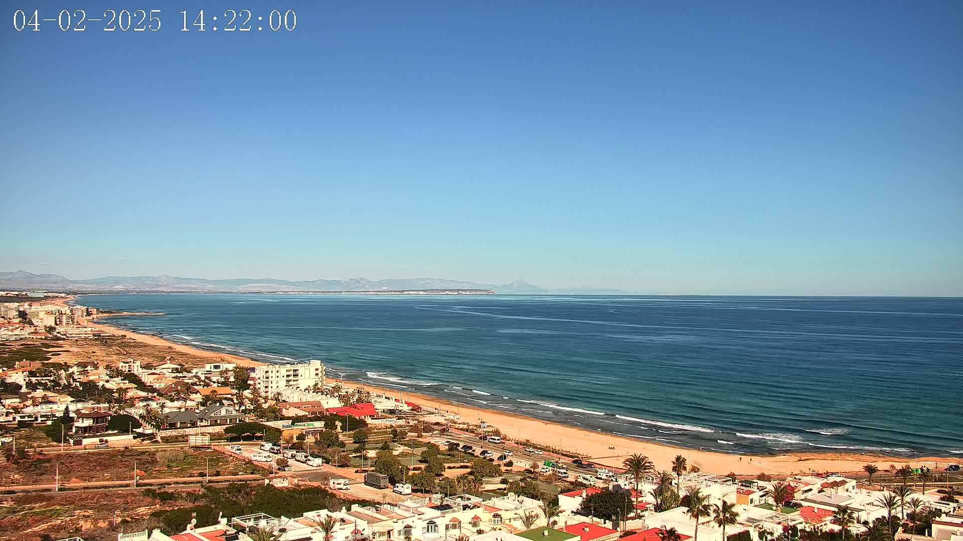 An expansive sandy beach with gentle waves stretches along a coastline, backed by a residential area under a clear blue sky and distant mountains.