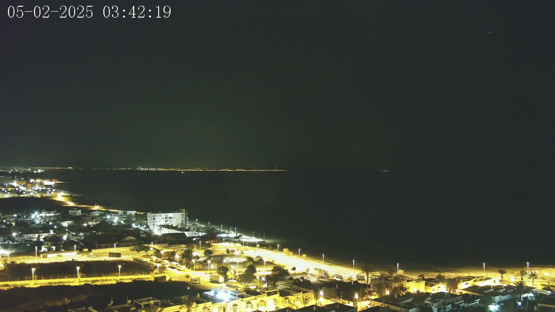 A nighttime coastal town with its lights illuminated against a dark, clear sky overlooks a calm, dark ocean.