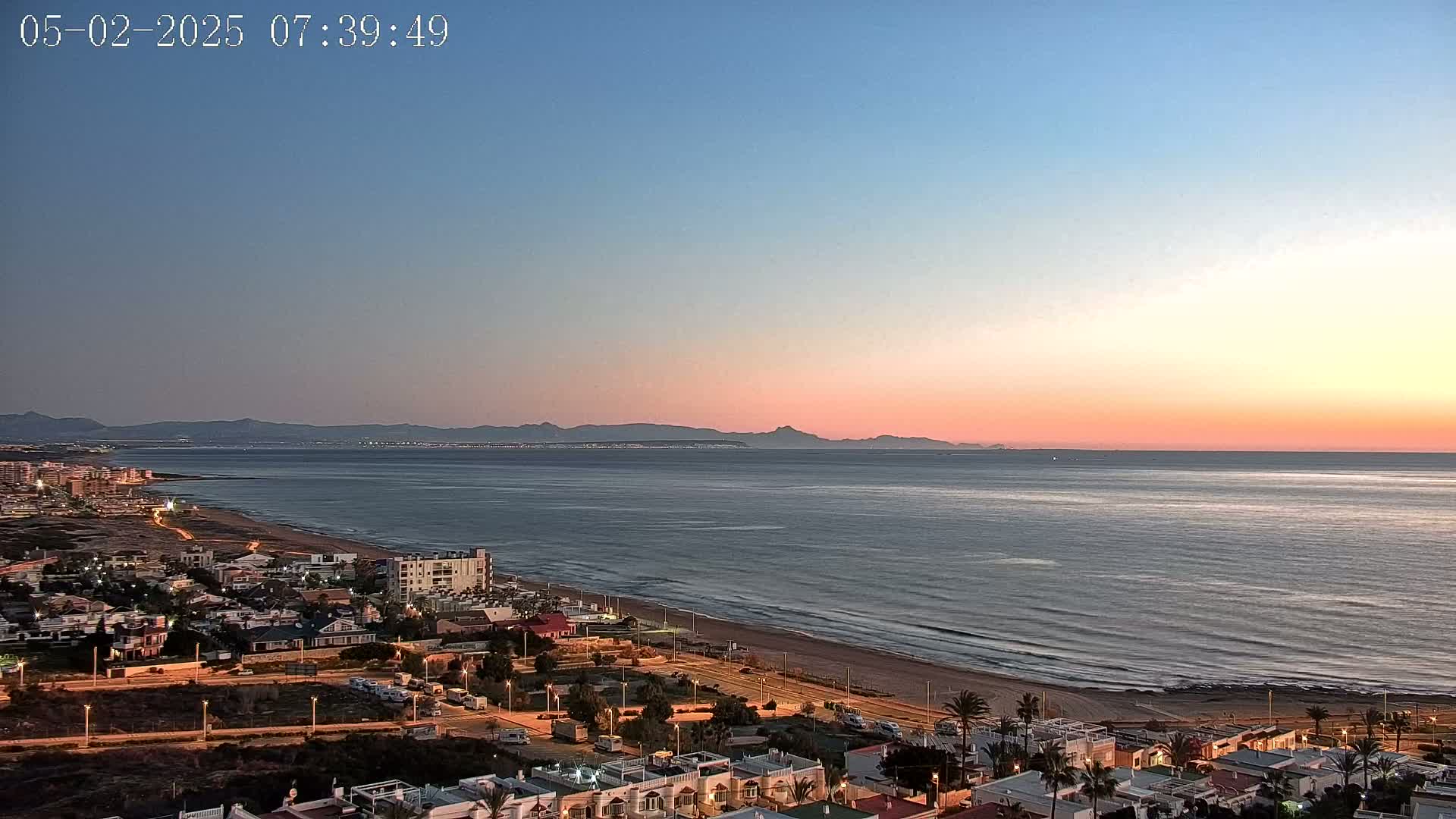A coastal town at dawn with a calm sea extending to distant mountains under a pink and blue sky.