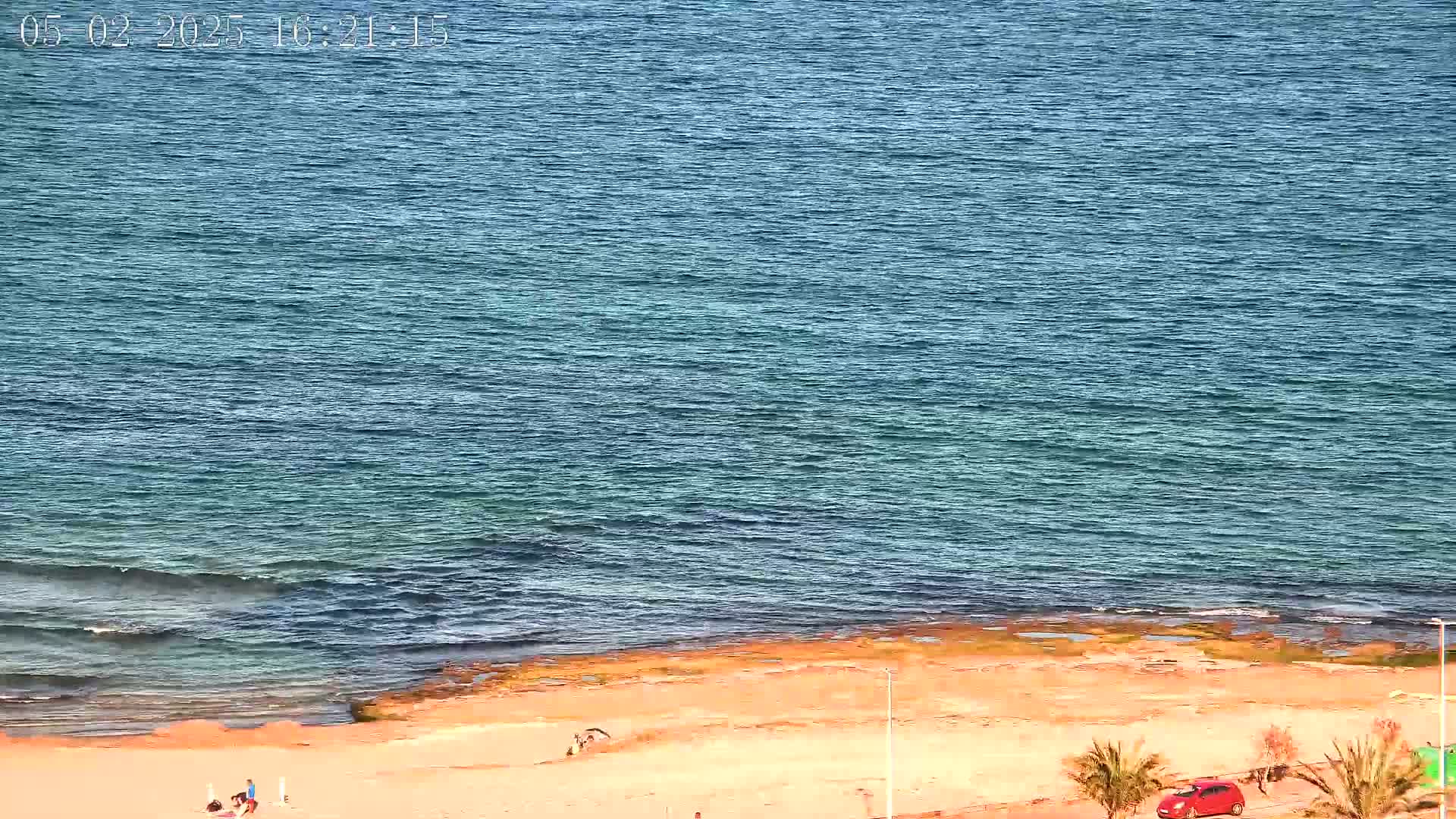 A sandy beach with a few people and a red car is next to a calm, blue ocean under a sunny sky.