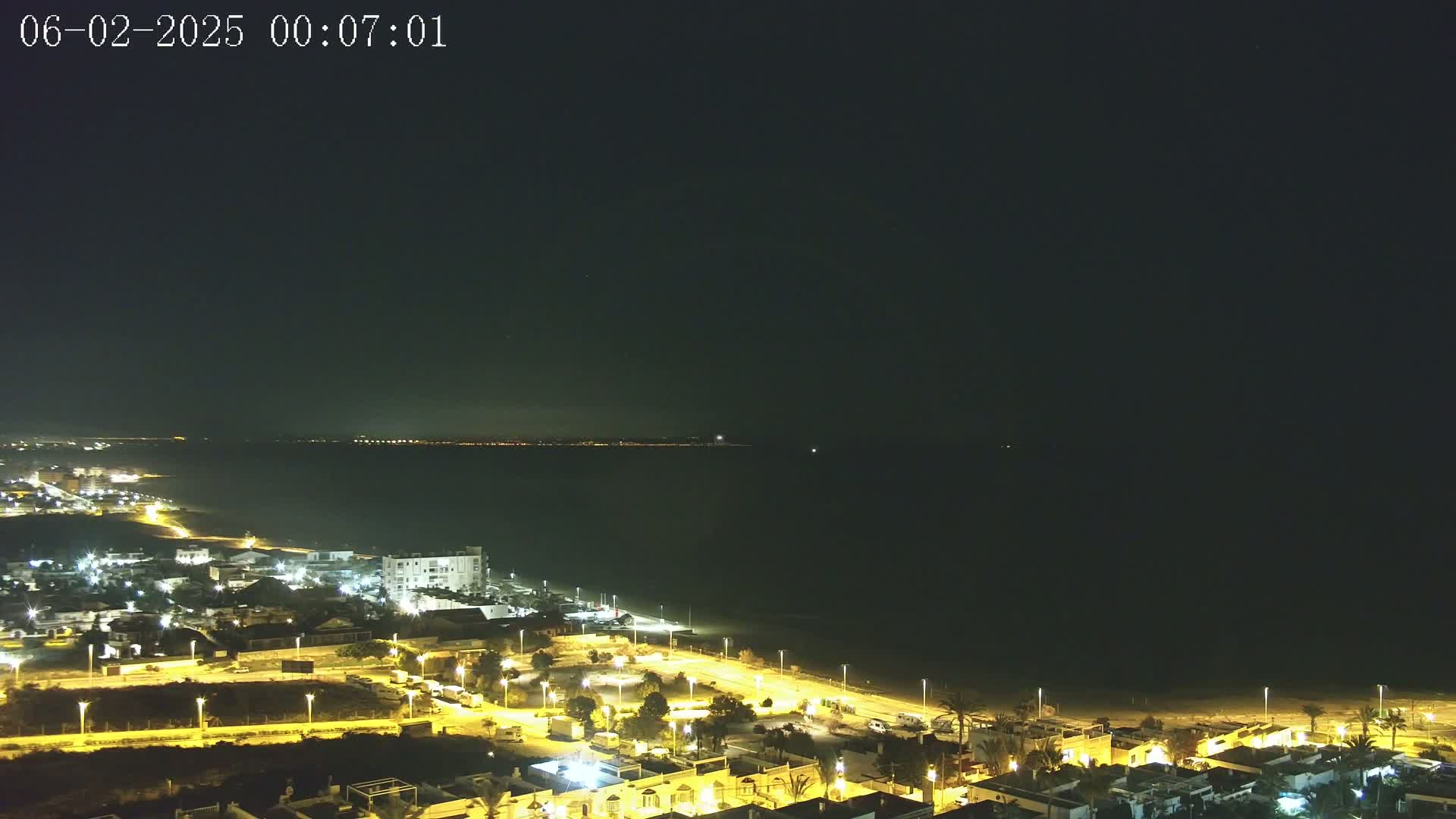 A nighttime coastal scene shows a dark body of water with a faintly lit distant shoreline, adjacent to a brightly lit town with streetlights illuminating buildings and roads.