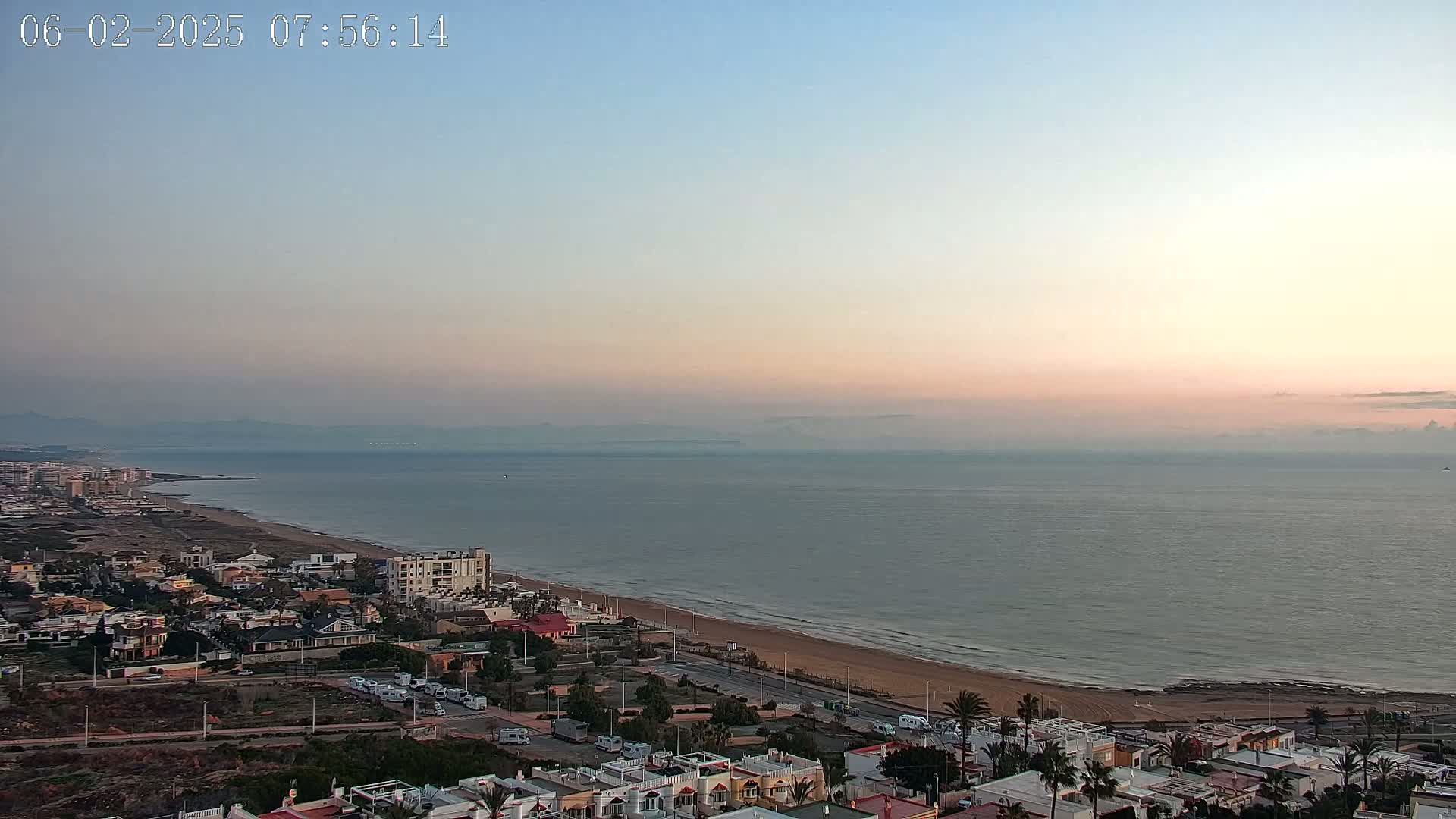 A coastal town at sunrise with a hazy, calm sea and a sandy beach.