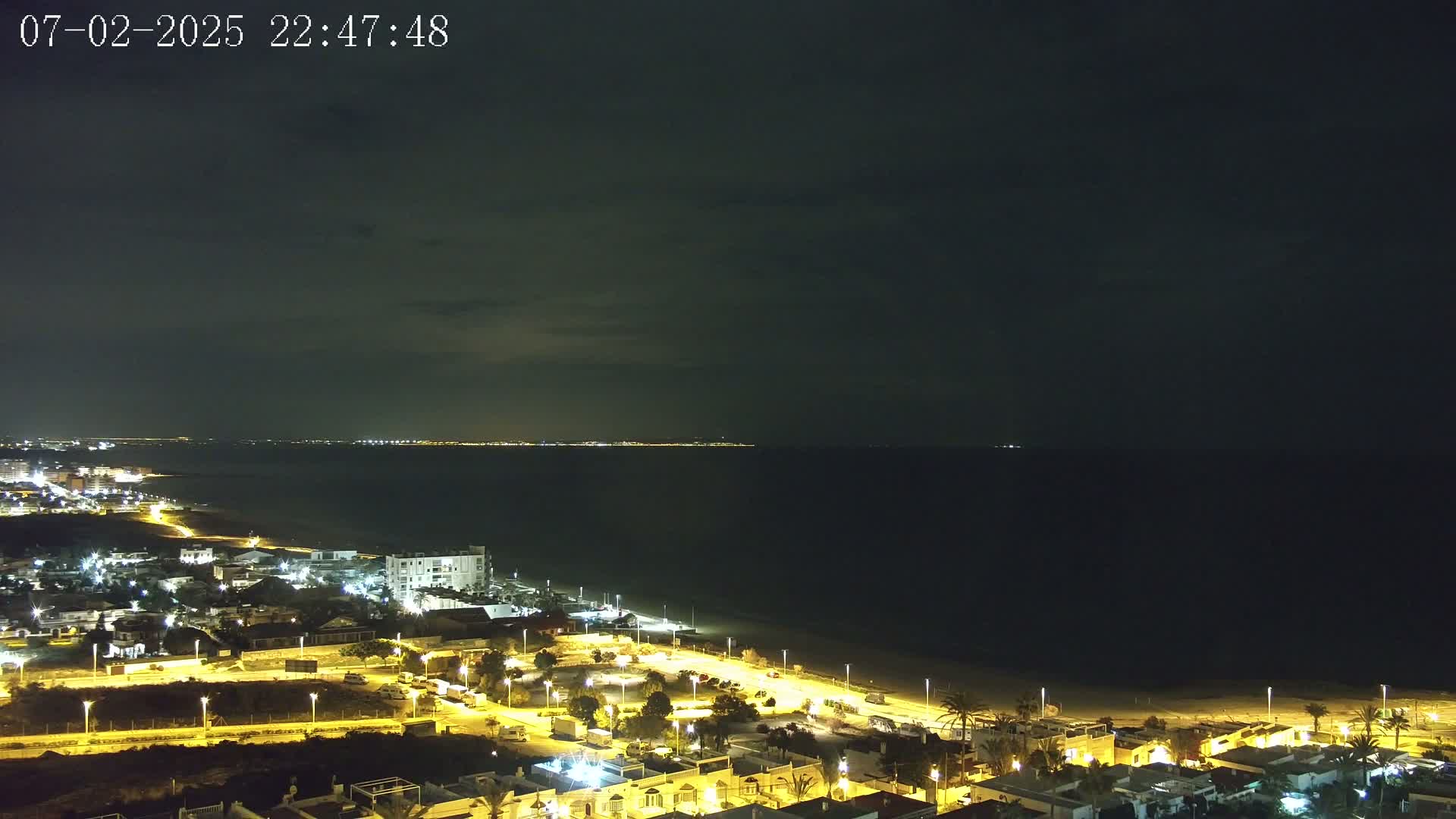 A nighttime coastal town is illuminated, with a dark ocean extending to a faintly lit horizon under a mostly clear, dark sky.