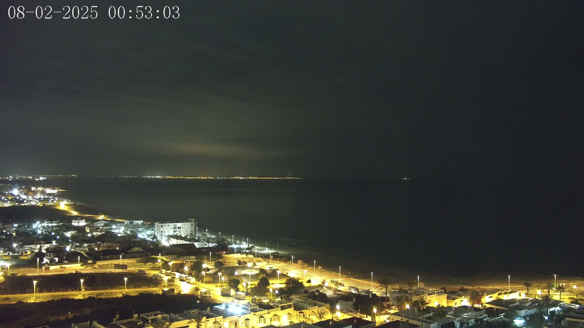 A nighttime coastal town with illuminated streets and buildings overlooks a dark ocean under a mostly clear sky.