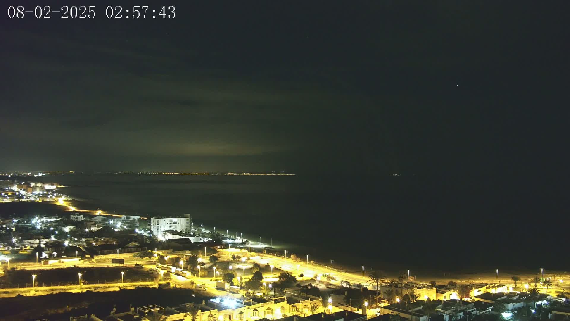 A nighttime coastal town is illuminated, with a dark ocean stretching to a faintly lit distant shoreline under a mostly clear sky.