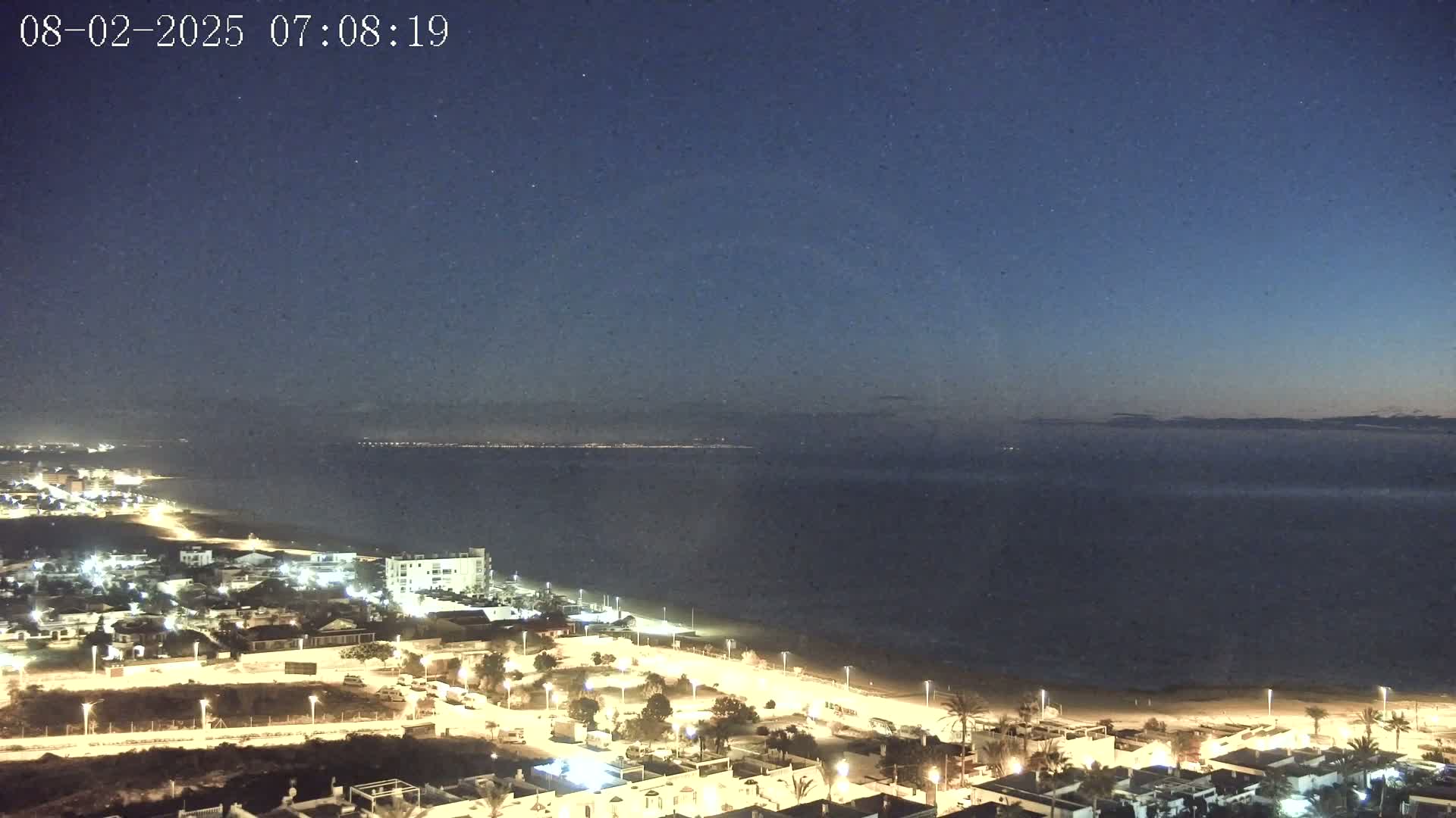 A nighttime coastal scene shows a dark ocean under a clear, starlit sky, with a town's illuminated buildings and streets bordering a sandy beach.
