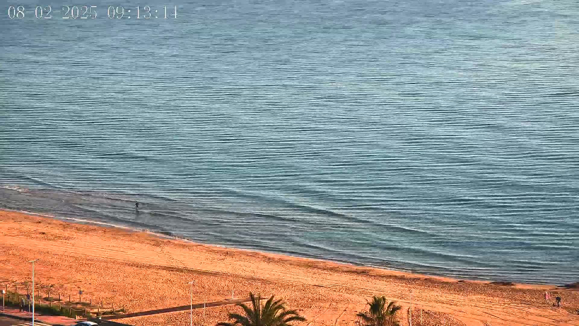 An aerial view shows a sandy beach meeting a calm, teal ocean under a clear sky.