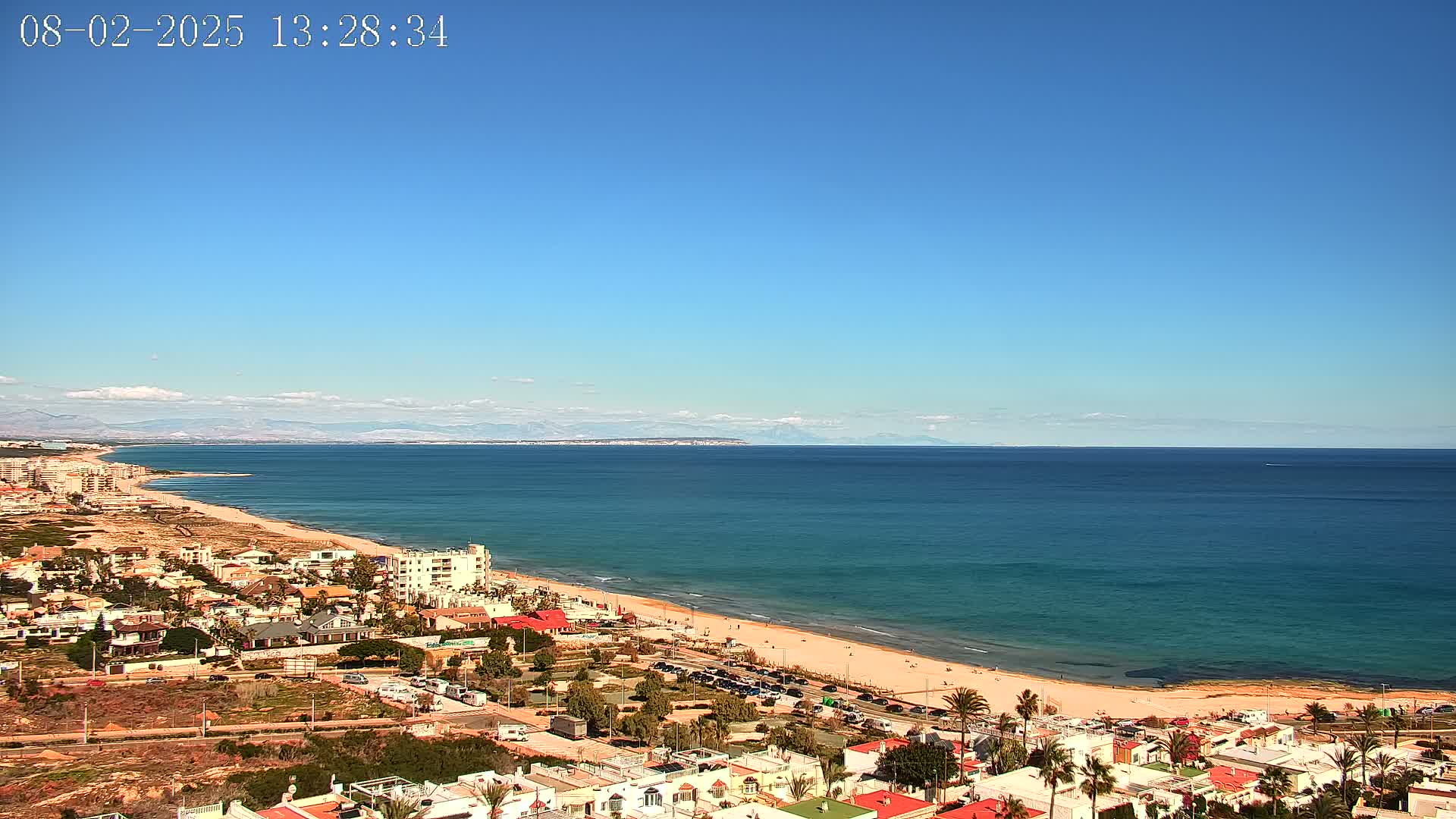 A mostly sunny coastal town overlooks a long sandy beach and calm blue ocean, with mountains visible in the far distance.