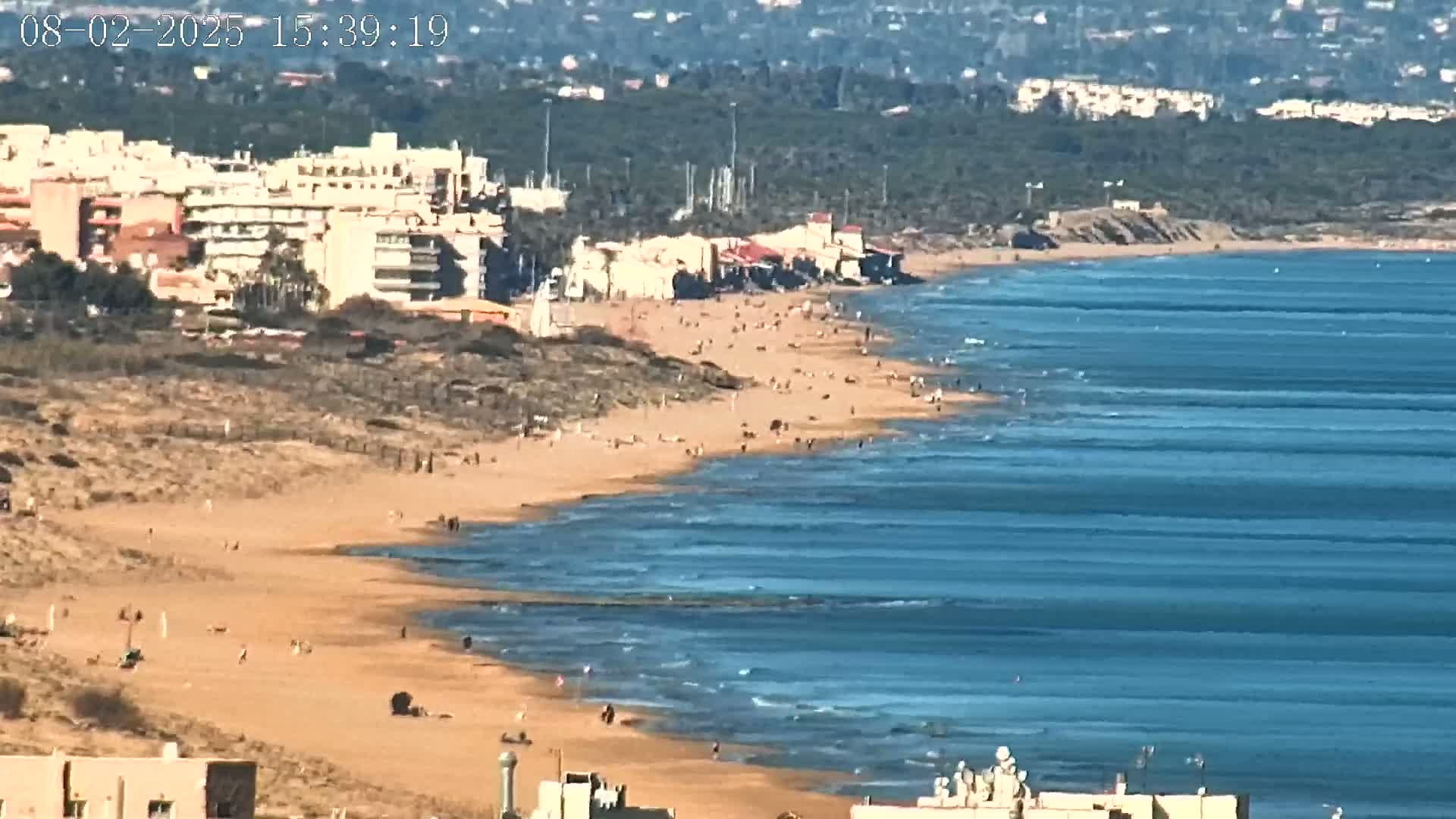 A long sandy beach with scattered people stretches along a calm, blue ocean, backed by buildings and a line of green vegetation under a clear sky.