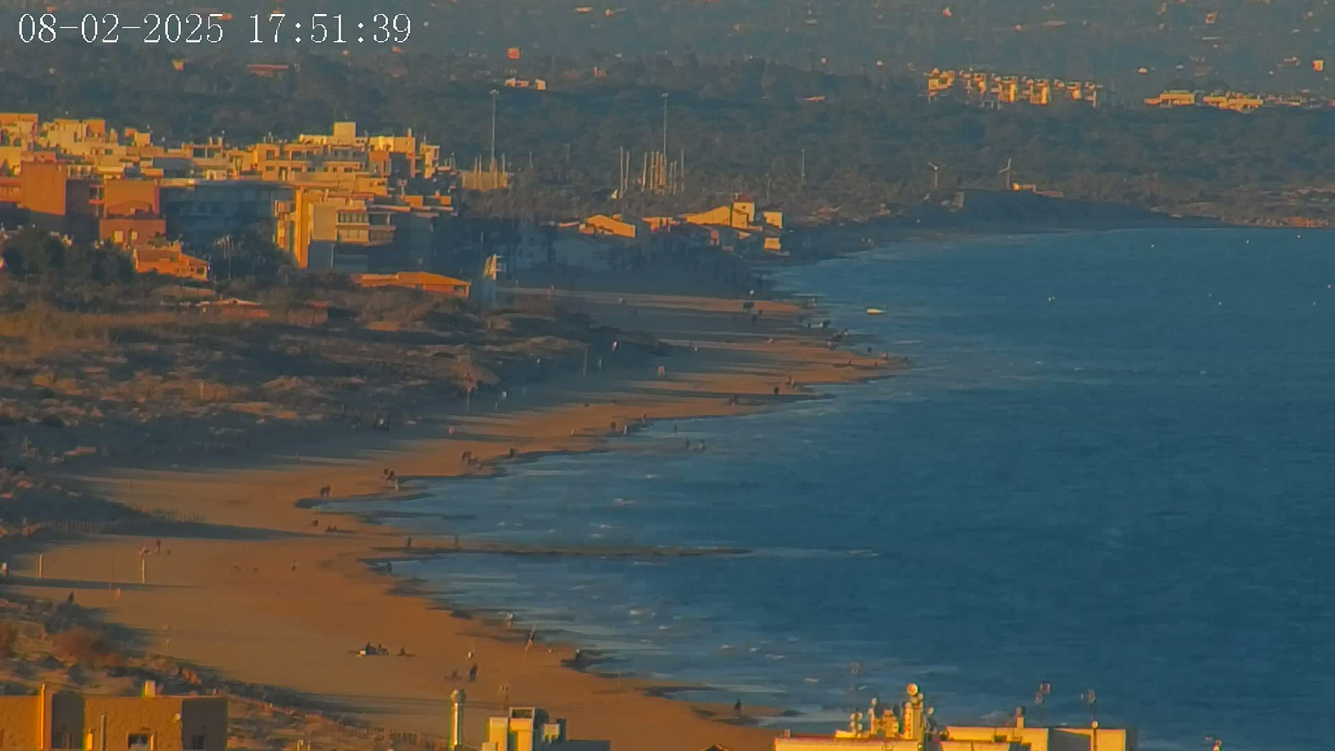 A sandy beach with sparse people stretches along a calm, blue-green sea under a clear, sunset sky, backed by a town of low-rise buildings.
