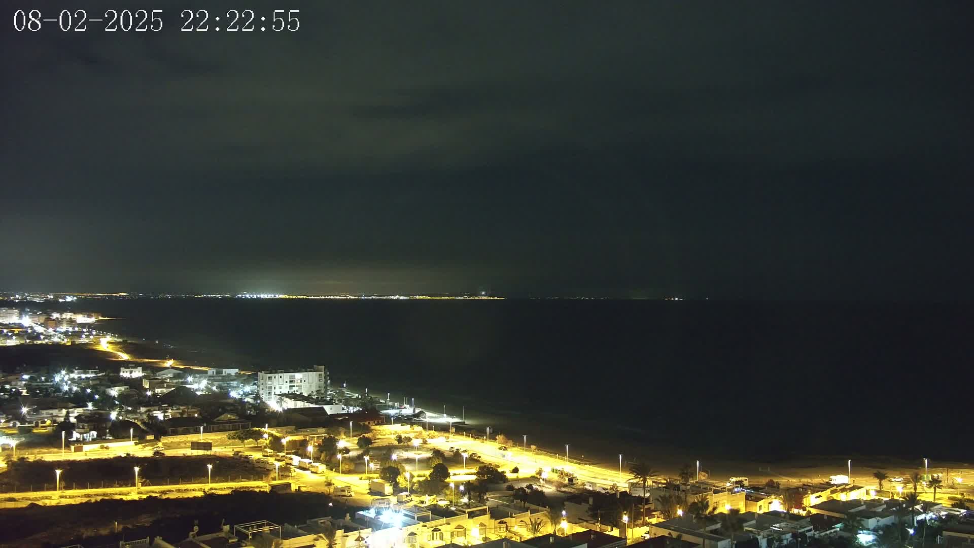 A nighttime coastal view shows a dark sea under a mostly cloudy sky, with a brightly lit town stretching along a sandy beach.
