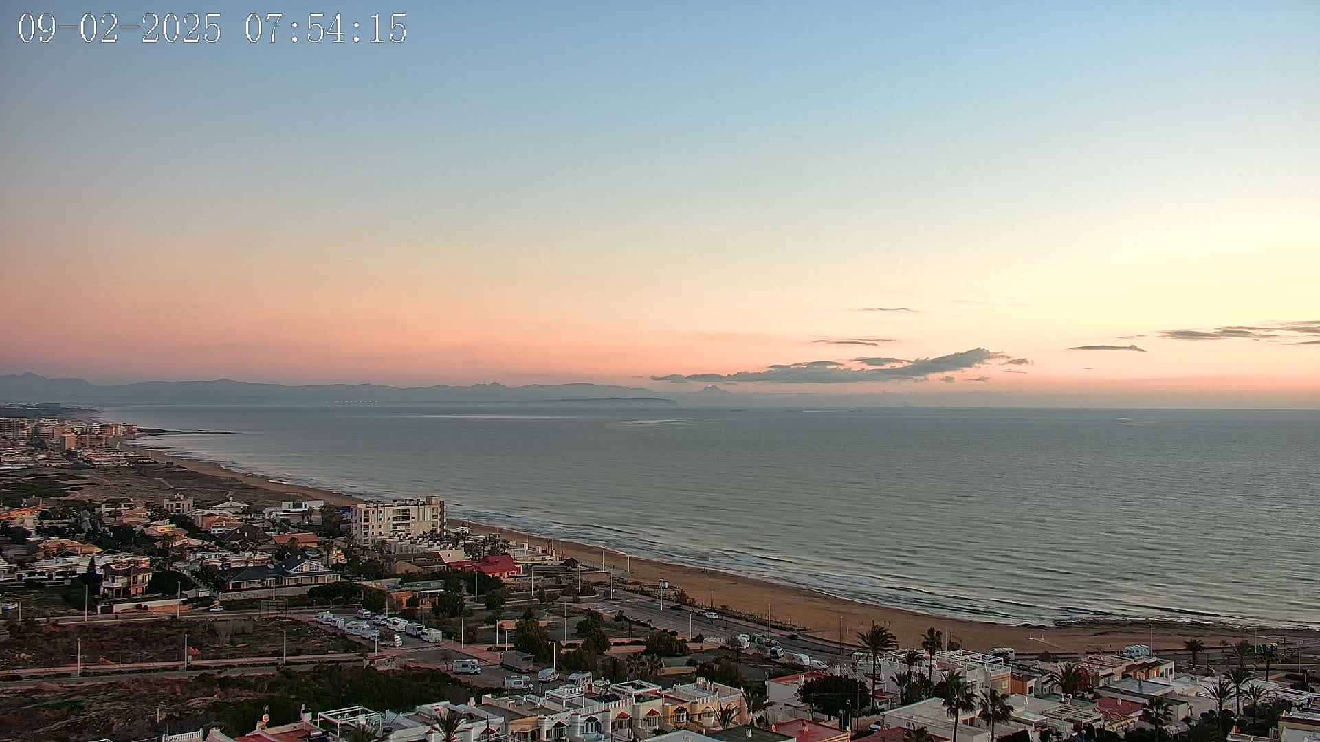 A coastal town at sunrise, with a calm sea and a pink and orange sky, overlooks a long sandy beach and distant mountains.