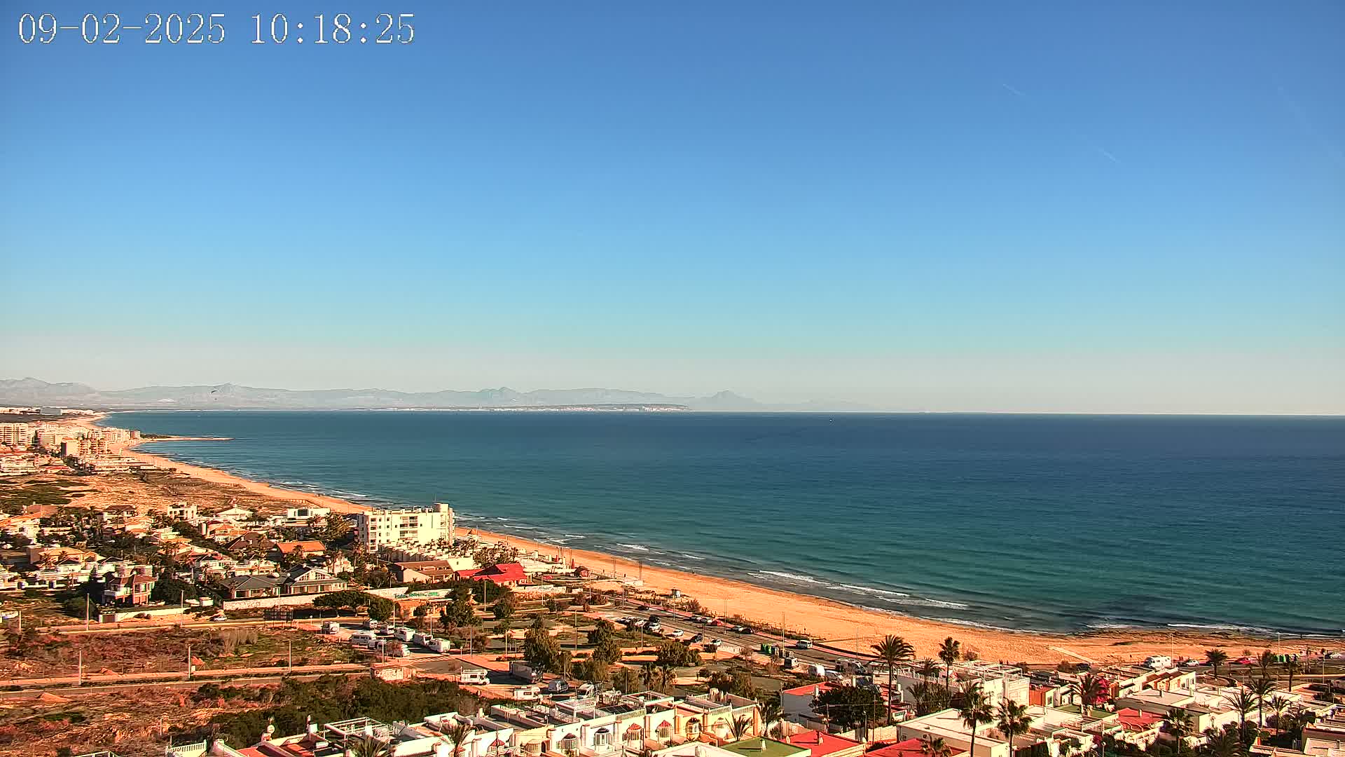 An expansive sandy beach borders a calm, teal ocean under a clear blue sky, with a residential area and distant mountains visible in the background.