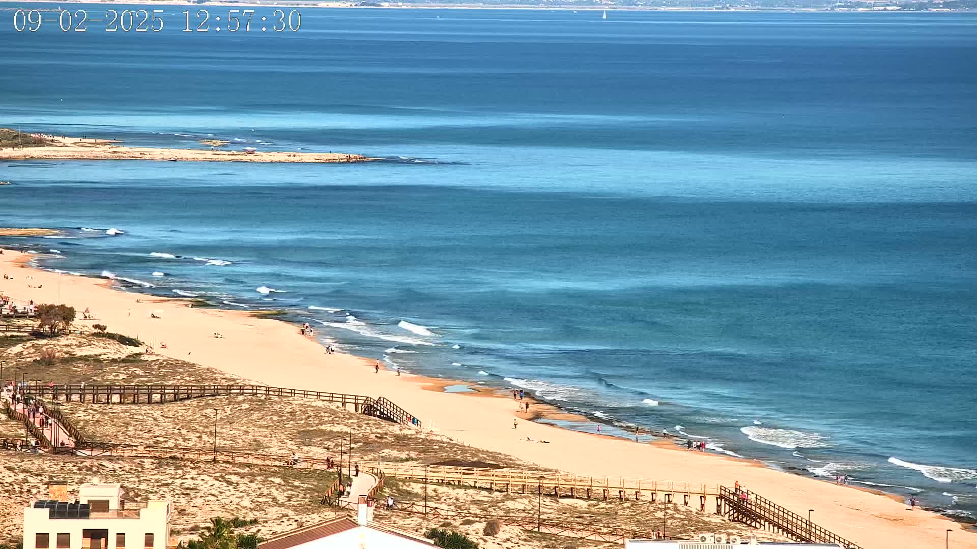 A sandy beach with people and wooden walkways is bordered by a calm, blue ocean with small waves under a sunny sky.