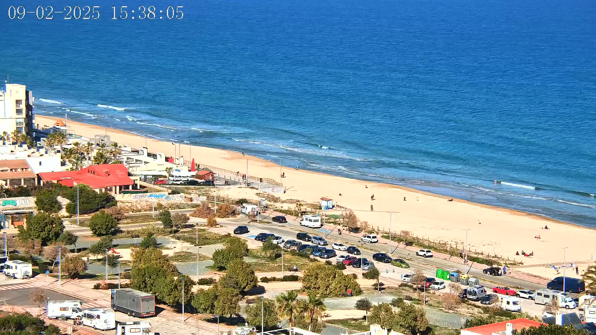 An aerial view shows a sandy beach with a few people on it, next to a parking lot filled with cars and RVs, all under a clear, sunny sky.