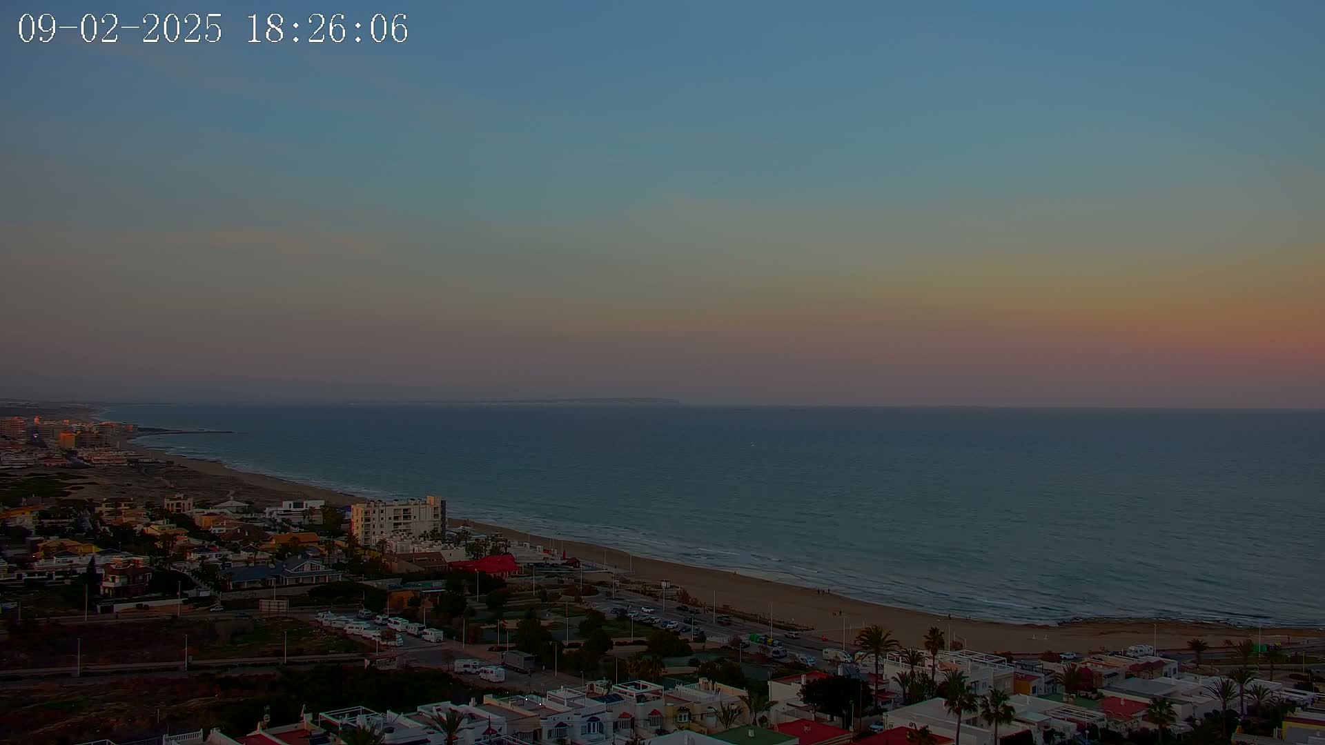 A coastal town at sunset, with a calm sea under a pale pink and blue sky.