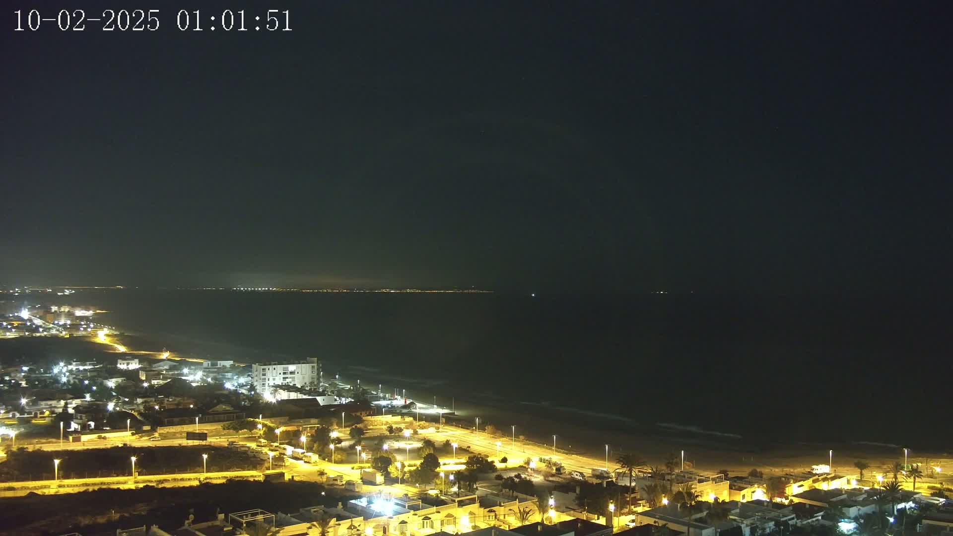 A nighttime coastal scene shows a dark ocean with gently lapping waves, a sandy beach, and a town illuminated by streetlights.