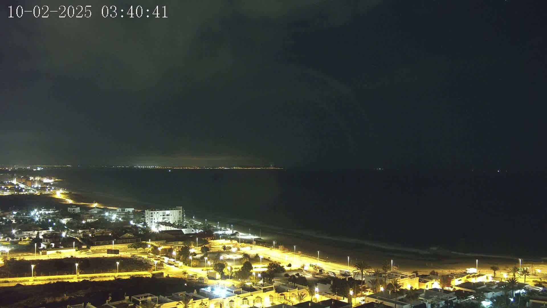 A nighttime coastal scene shows a dark ocean with gently lapping waves on a sandy beach, and a town with illuminated buildings and streets stretching inland under a mostly cloudy sky.