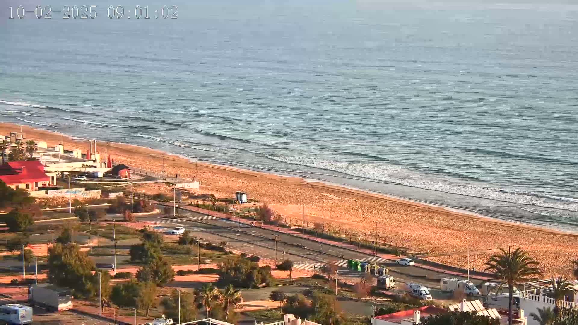 An aerial view shows a sandy beach with gentle waves lapping the shore on a partly sunny day, with a road and buildings visible beyond.