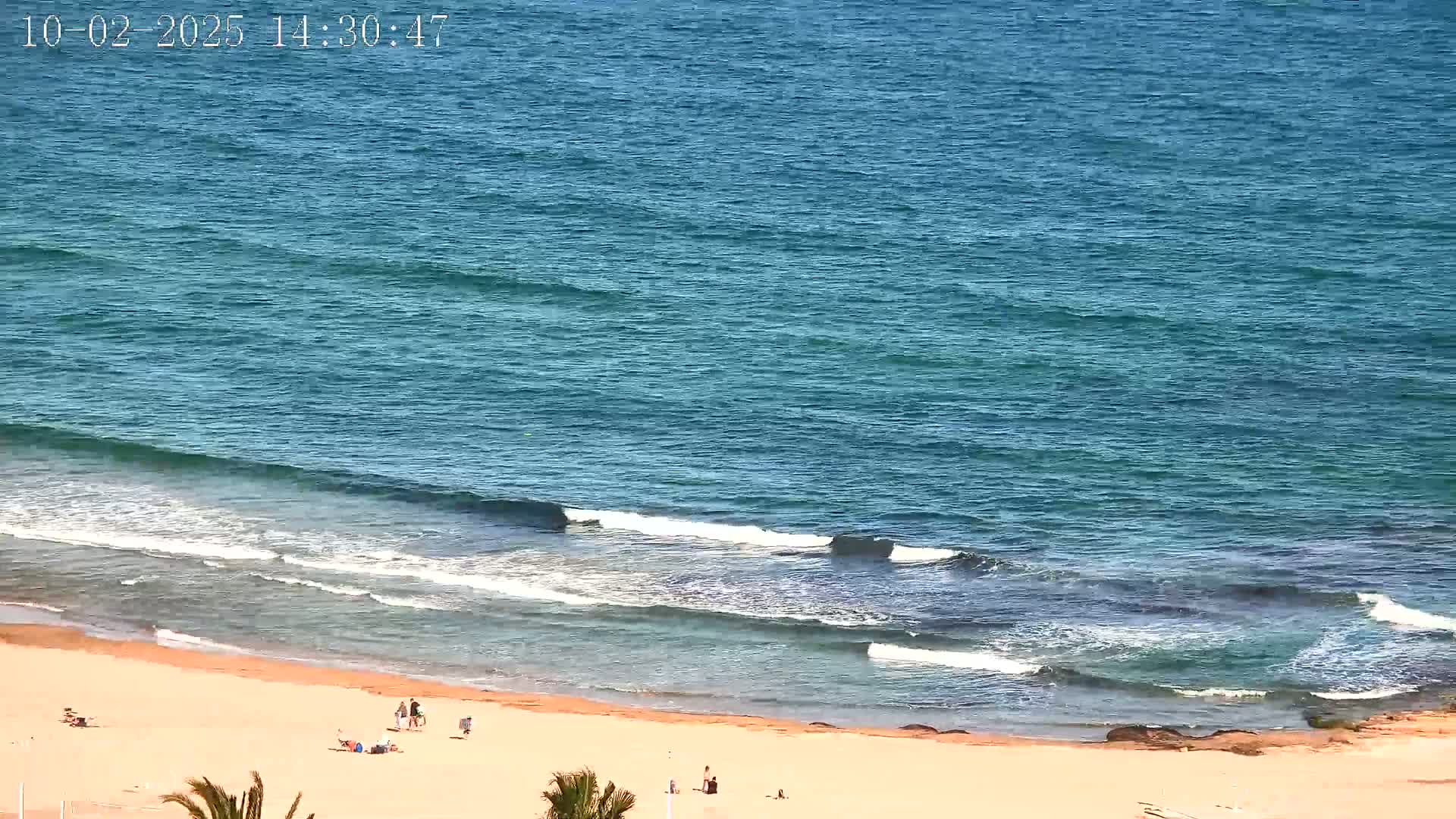A sandy beach with a few people on it is gently lapped by small waves on a clear, sunny day.