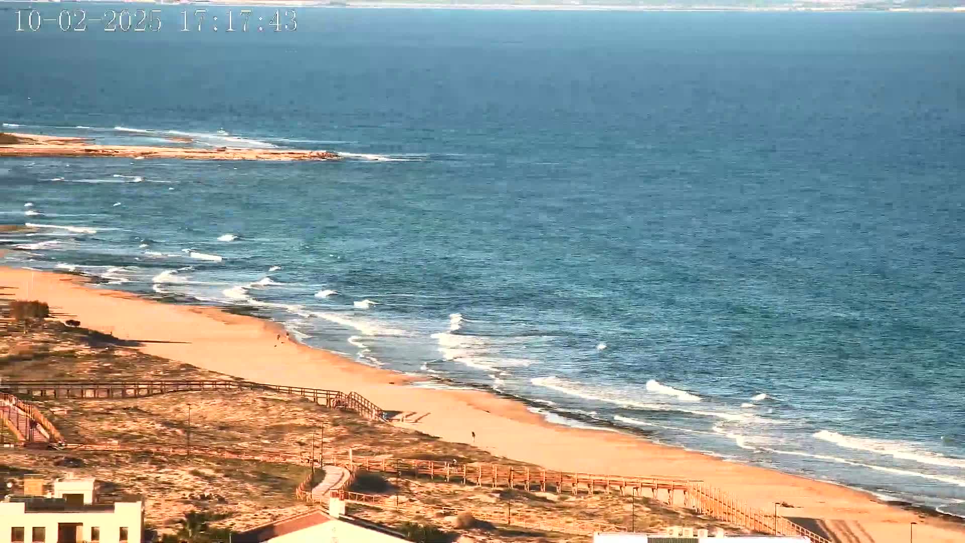 An aerial view shows a sandy beach with gentle waves lapping the shore under a clear sky.