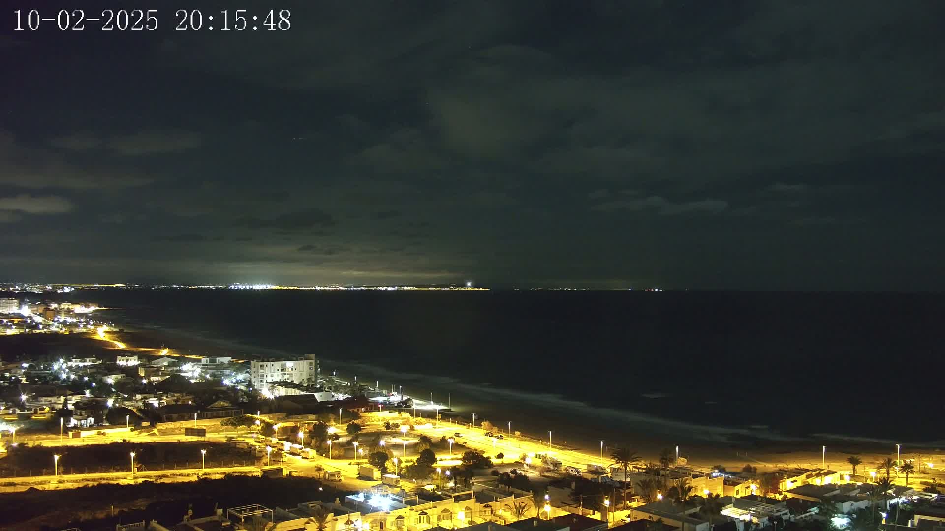 A nighttime coastal scene shows a dark ocean under a cloudy sky, with a town's lights illuminating the beachfront and nearby streets.