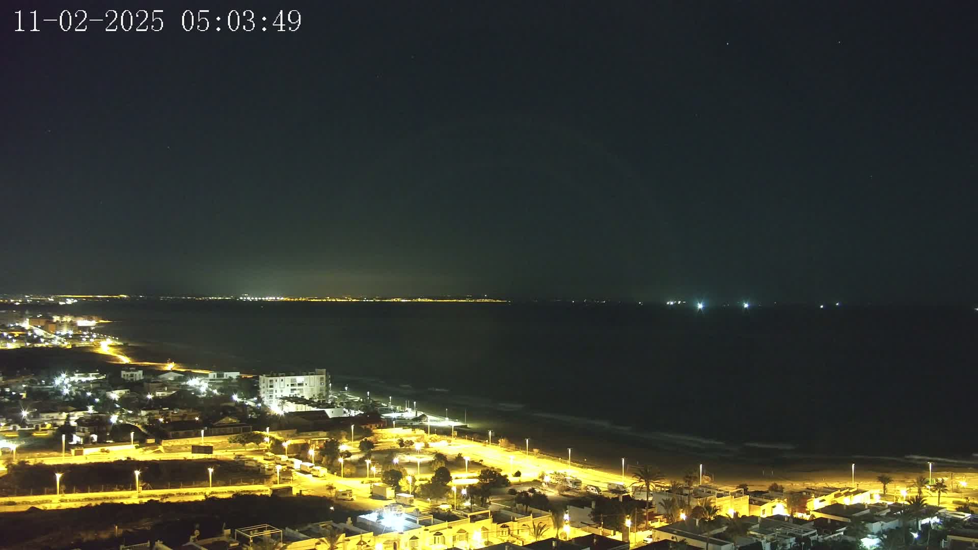 A nighttime coastal scene shows a dark ocean with small waves lapping a sandy beach, bordered by a town with illuminated streets and buildings under a clear, starlit sky.