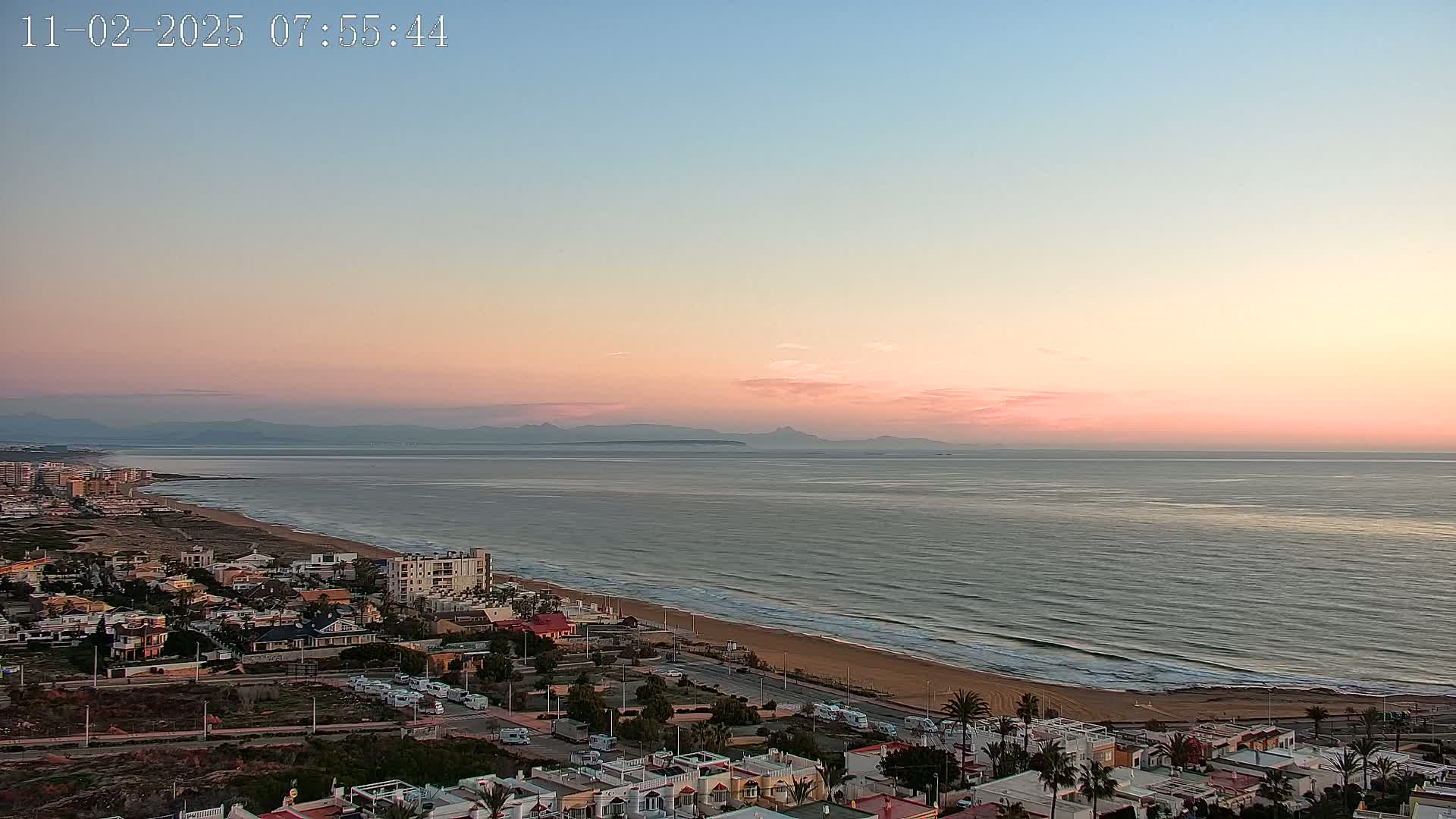 A coastal town at sunrise overlooks a calm ocean with a sandy beach, under a pale pink and blue sky.